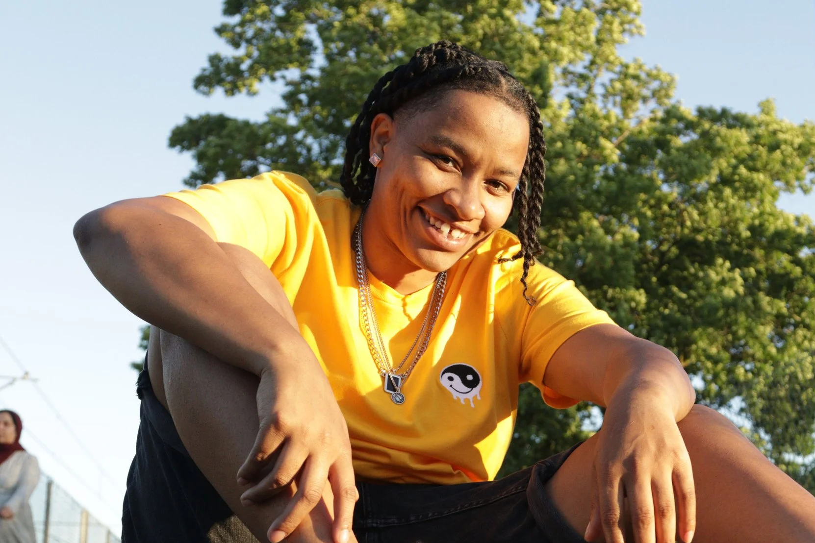 A young woman sitting on a running track, smiling, wearing a yellow t-shirt, black shorts, white sneakers, and jewelry, with greenery and a chain-link fence in the background.