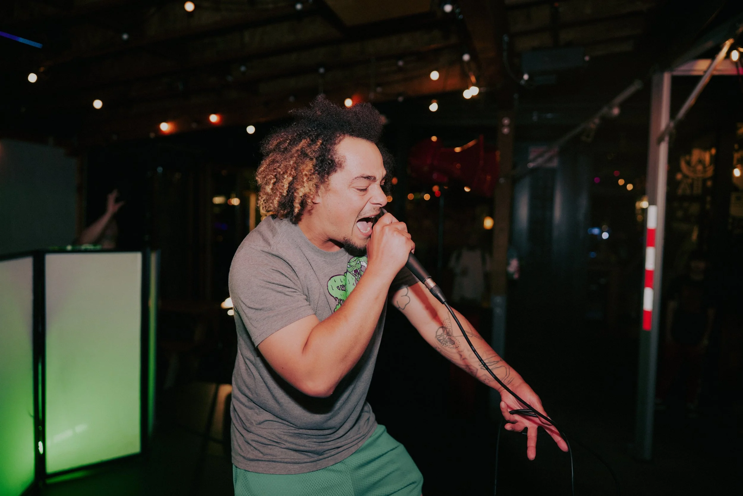 A man with curly hair singing into a microphone at a karaoke bar or stage, with a DJ booth in the background.