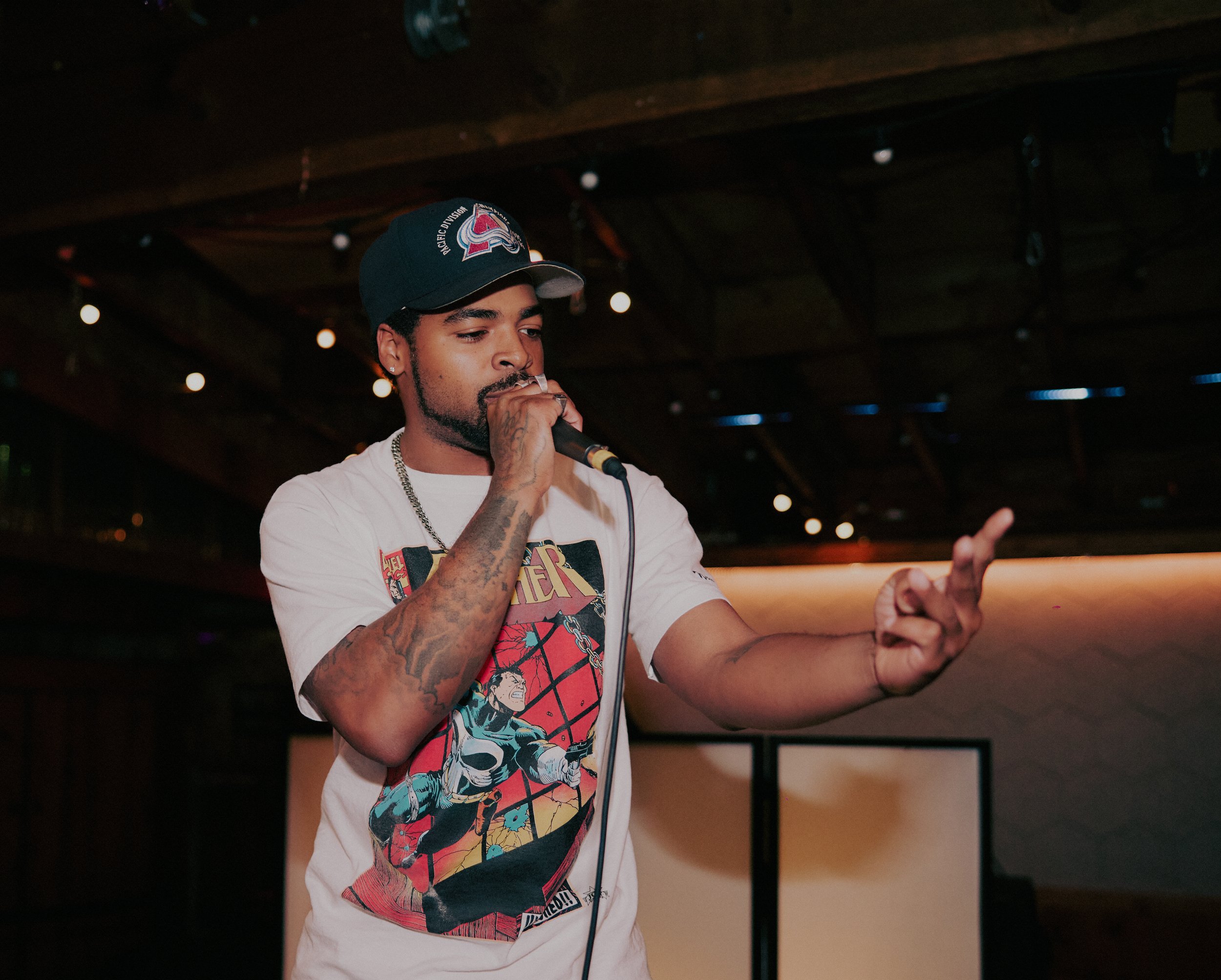 A young man performing with a microphone in a dimly lit indoor venue, wearing a Black Colorado Avalanche baseball cap and a white Marvel Thor T-shirt, with tattoos on his arms.
