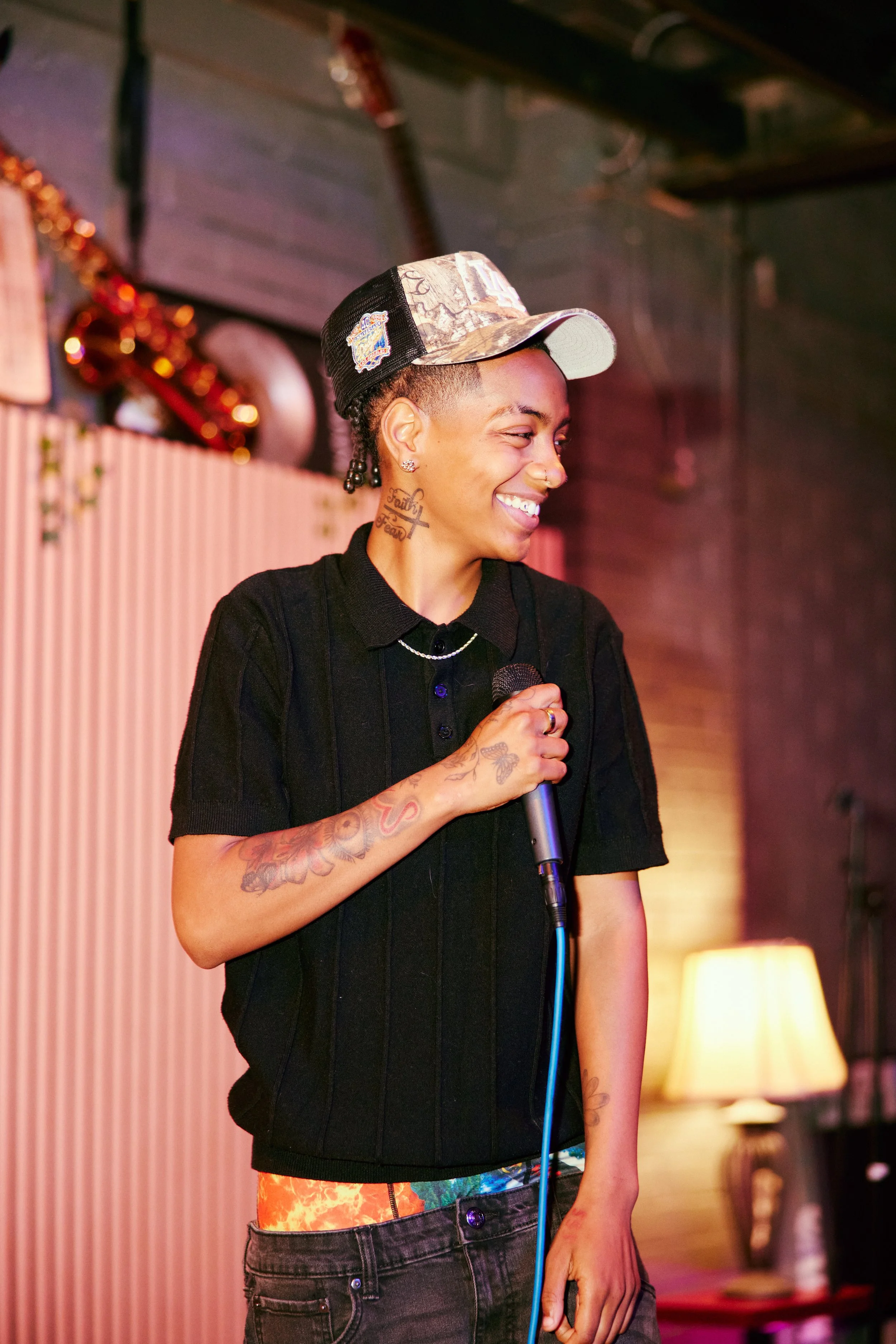 A woman with tattoos and earrings smiling while holding a microphone, wearing a black shirt and a camouflage baseball cap. She appears to be at a karaoke or open mic event.