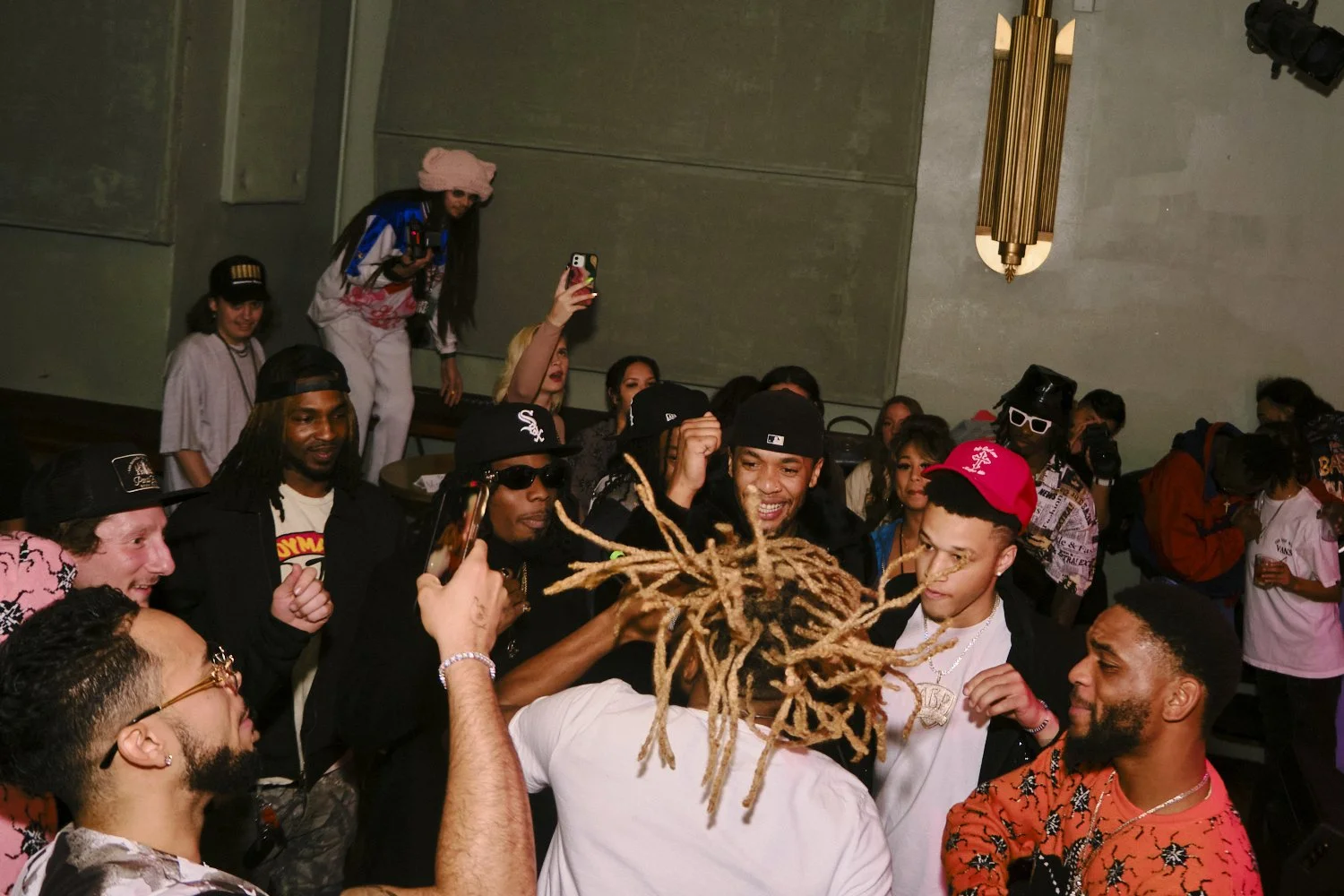 Group of people at a social event or party, some taking photos or videos, others smiling and talking, with a person with dreadlocks facing away from the camera in the foreground.