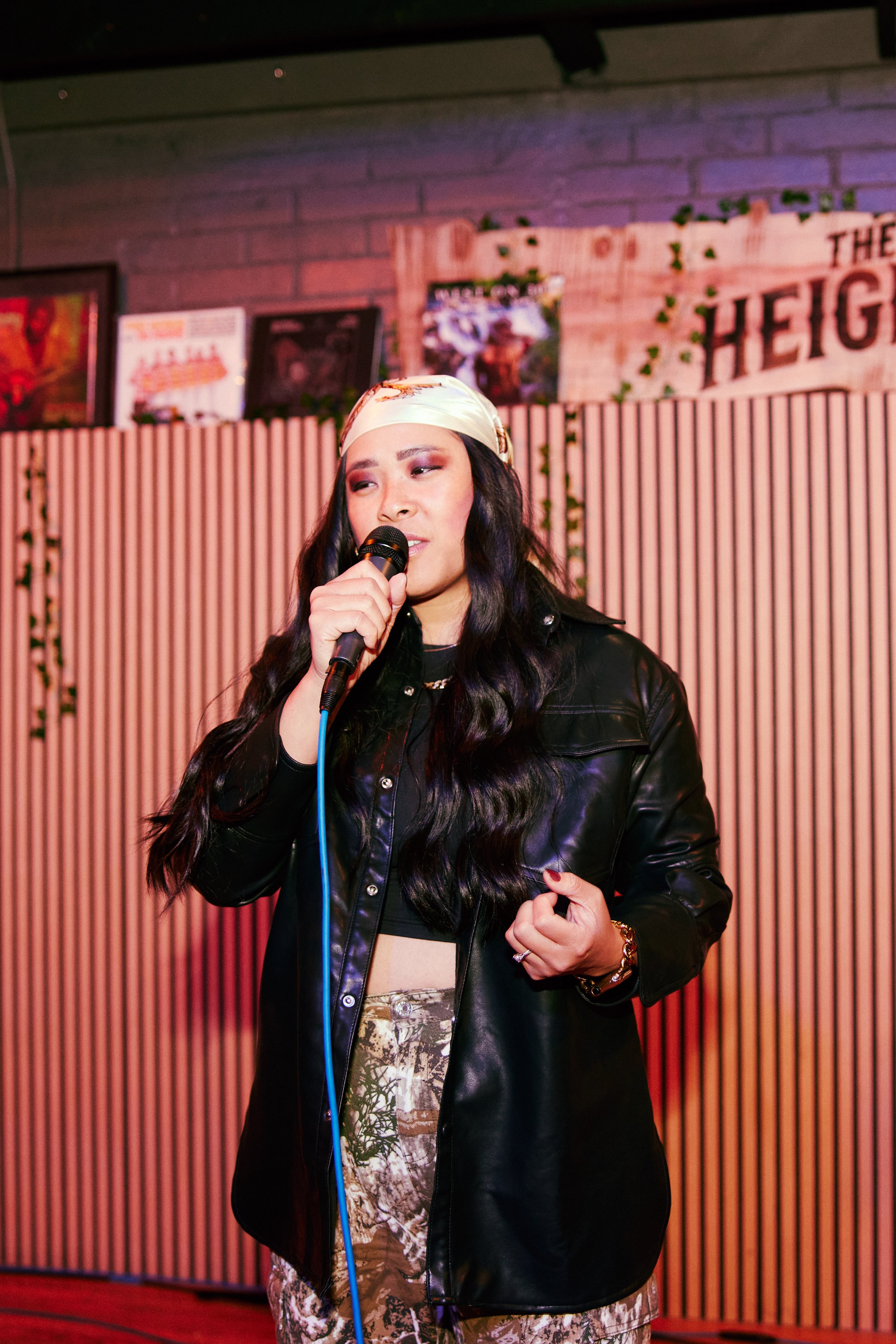 A woman with long dark hair singing into a microphone on stage, wearing a black leather jacket, patterned pants, and a white headscarf in a venue with a wooden wall backdrop.
