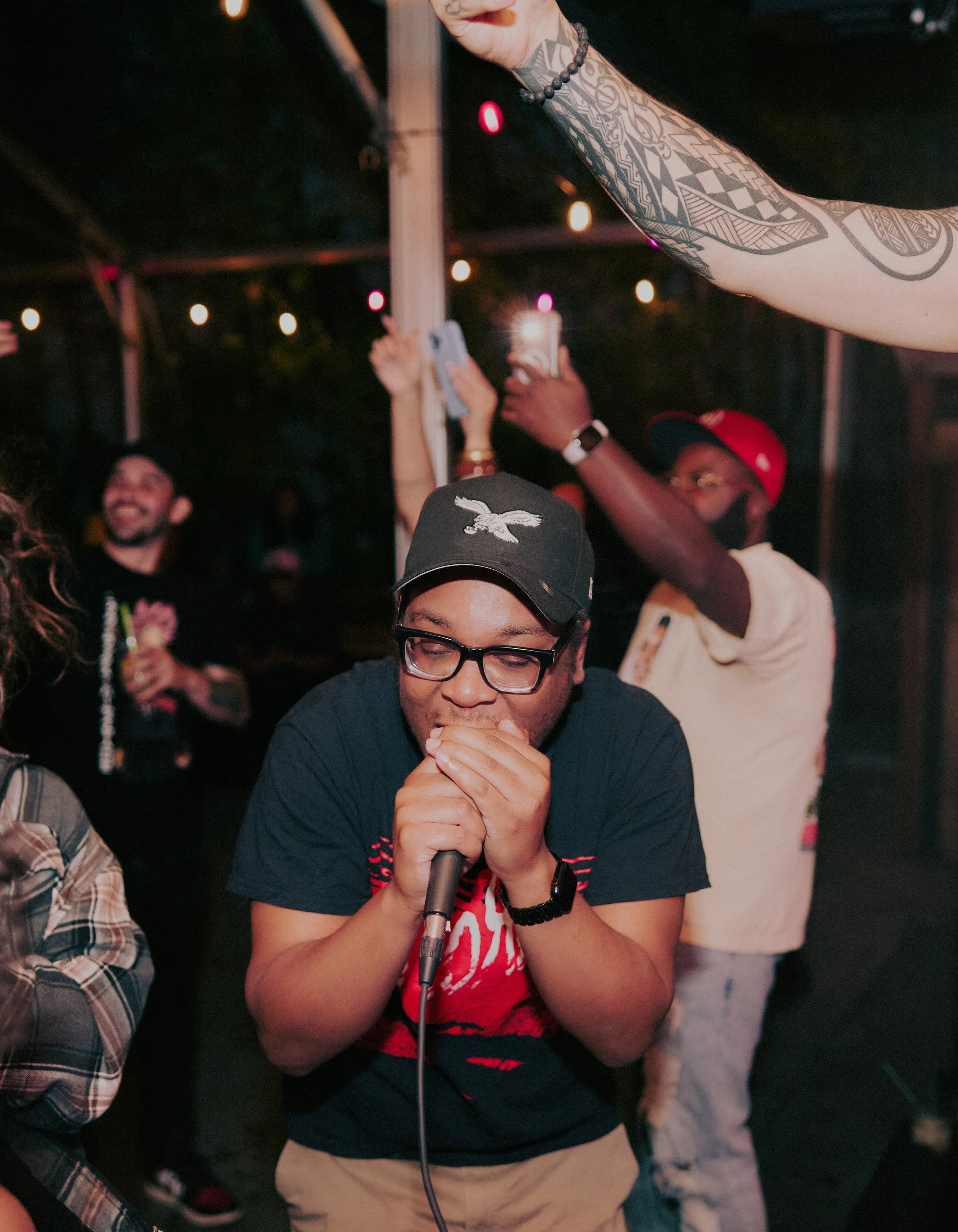 A young man holding a microphone sings or speaks at a lively outdoor gathering at night, with people around him taking photos and enjoying the event, under string lights.