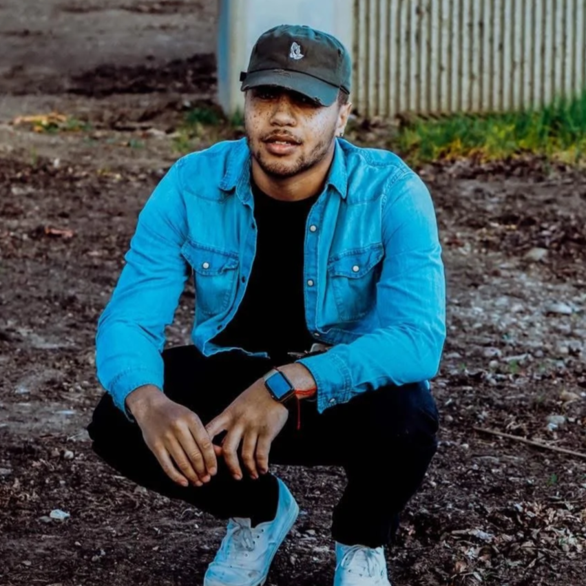 A young man squatting outdoors on dirt, wearing a black cap, blue denim jacket, black pants, white sneakers, and a smartwatch. There are concrete structures and a wooden fence in the background.