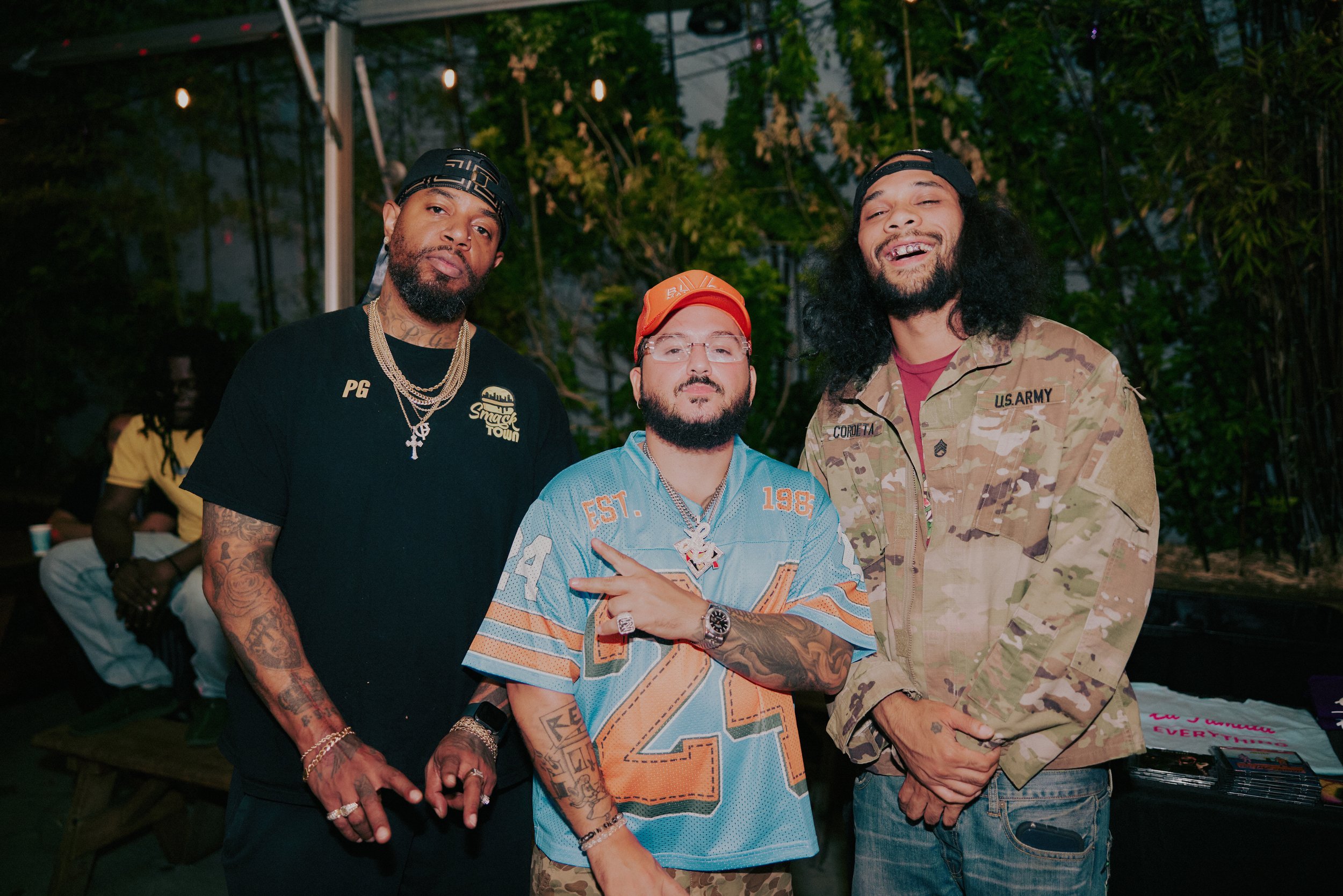 Three men standing close together outdoors at night, in front of string lights and greenery, posing for the photo. The man on the left is wearing a black t-shirt with gold chains, and has tattoos on his arms. The man in the middle is wearing glasses, a bright orange cap, a sports jersey, and has tattoos on his arms, displaying a hand gesture. The man on the right is wearing a camouflage U.S. Army jacket and a backwards cap, with long curly hair and a beard.