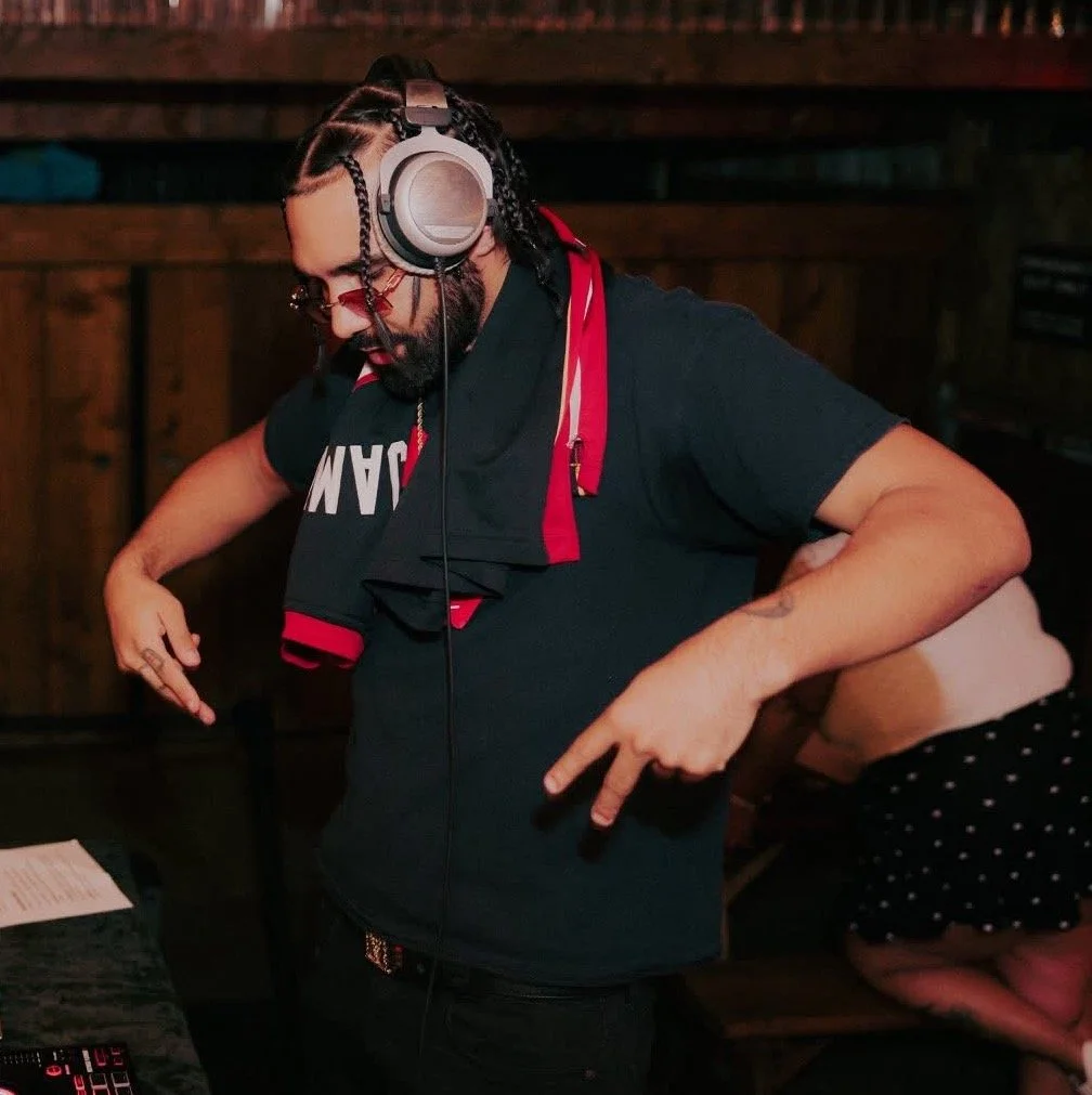 A person with braided hair wearing headphones, a black shirt with red and white accents, standing at a DJ controller with one hand on the controls, in a dimly lit indoor setting.