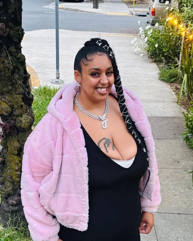 A woman smiling and winking at the camera sitting at a wooden table in a room with dark wood and brick walls, holding a pair of sunglasses, with flowers and miscellaneous items on the table.