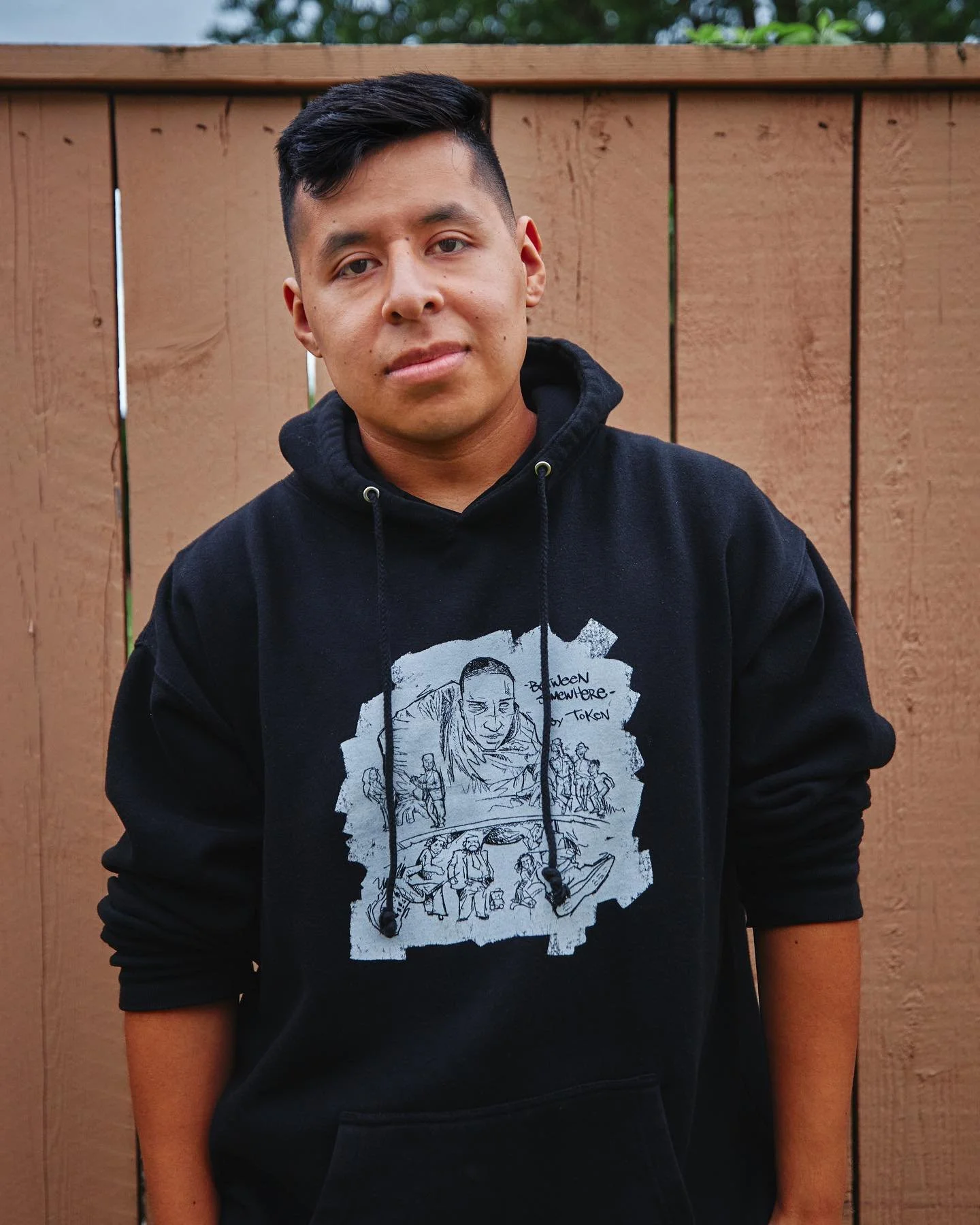 Young man standing outdoors in front of a wooden fence, wearing a black hoodie with a graphic design on the front.
