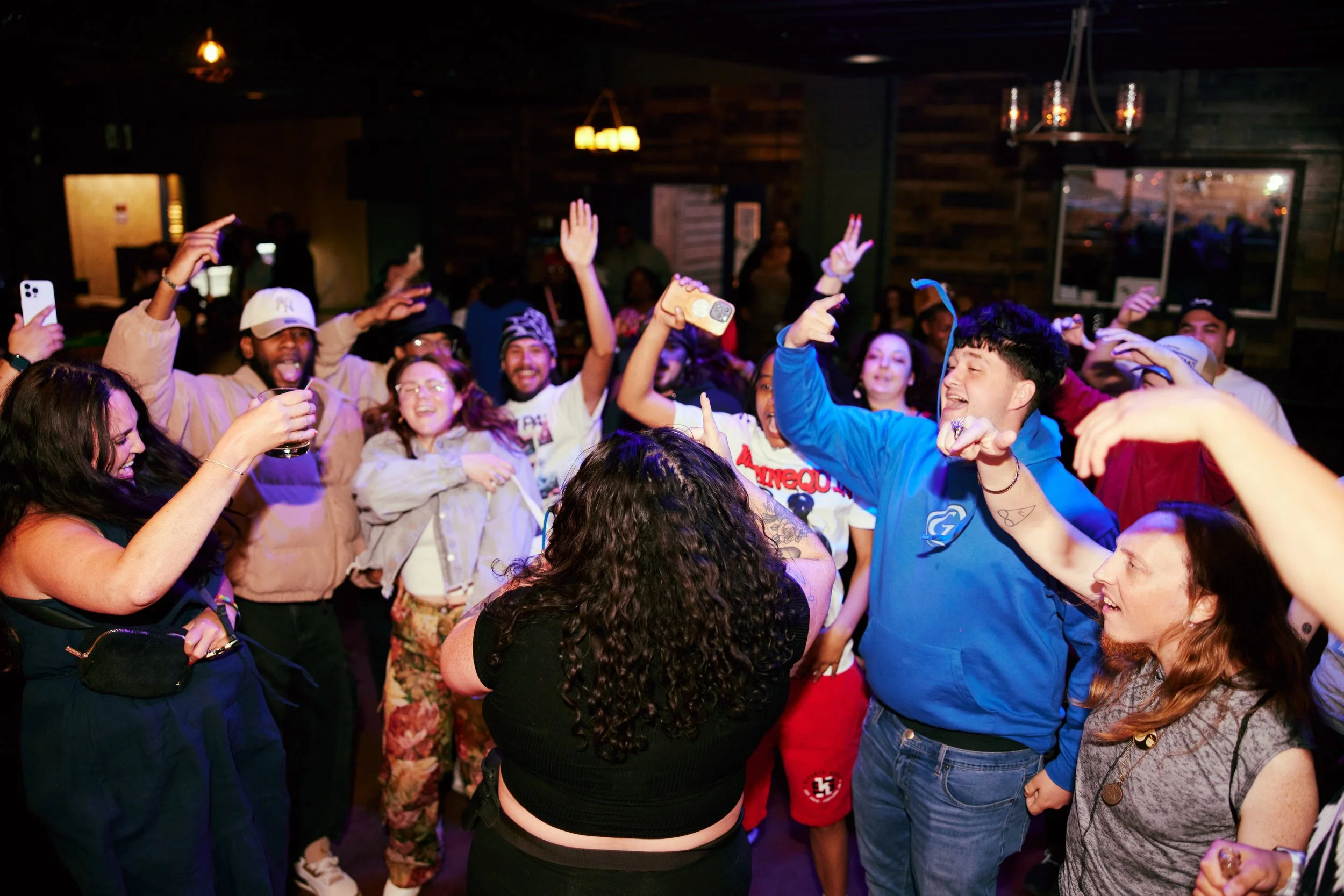 Group of people dancing and celebrating in a dimly lit indoor setting, some with raised hands, smiling, and holding drinks or phones.