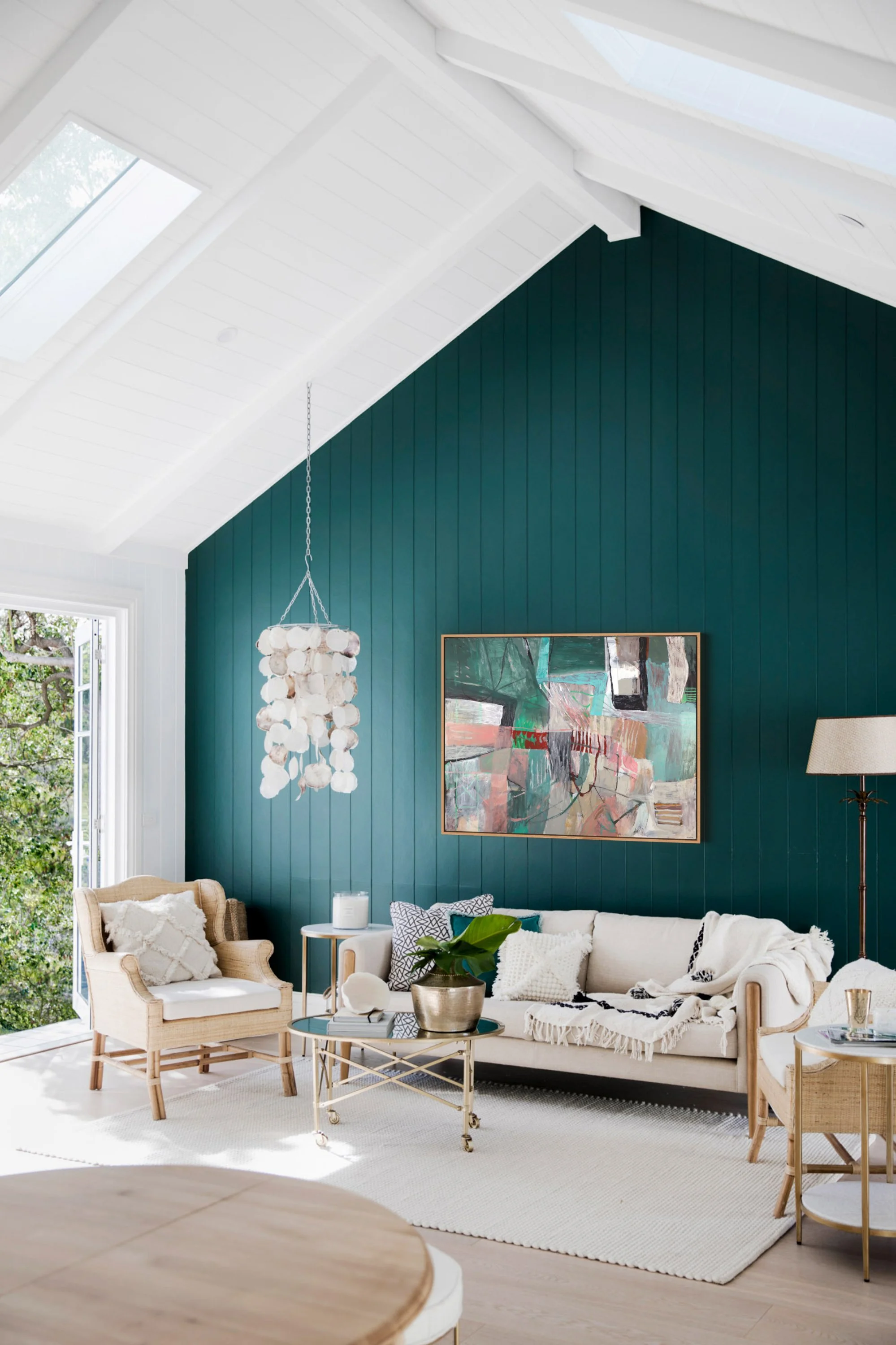 Bright living room with white and wood furniture, a sofa with pillows, a coffee table with a plant, and a large abstract painting on a teal wall. Large windows and skylights let in natural light.