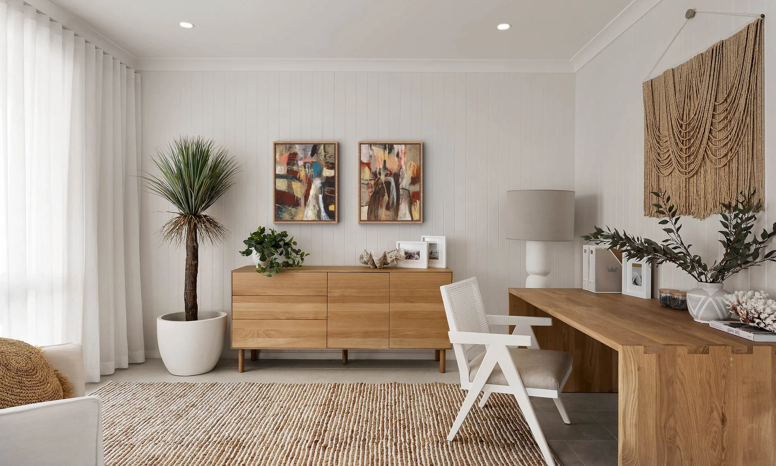 Interior of a modern living room with white walls, a wooden sideboard, abstract artwork, large potted plant, and a wooden dining table with white chairs.