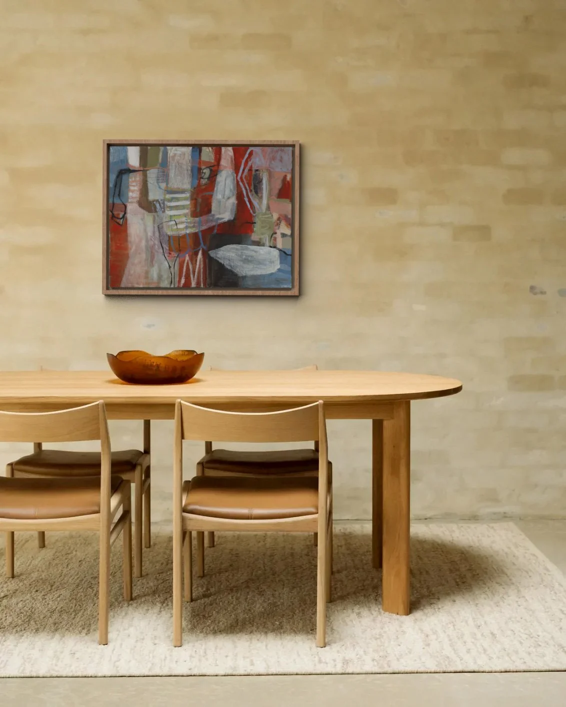 A wooden dining table with four matching chairs, a wooden bowl on the table, a beige textured wall with an abstract painting framed on it, and a beige rug underneath the table.