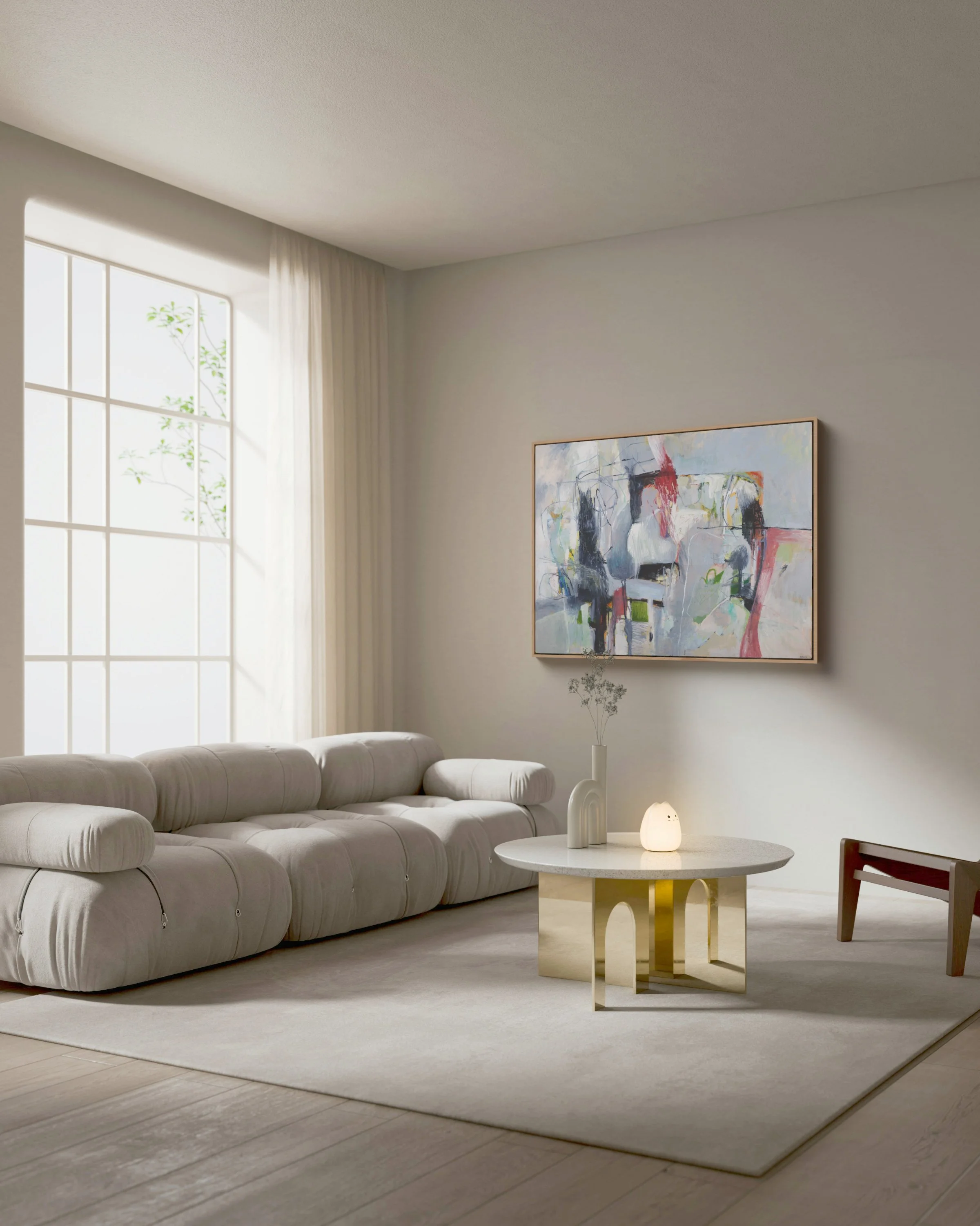 A cozy living room with a large beige sectional sofa, a round white coffee table with gold legs, a small side table with a decorative vase, a lamp, modern artwork on the wall, and a large window with sheer curtains letting in natural light.