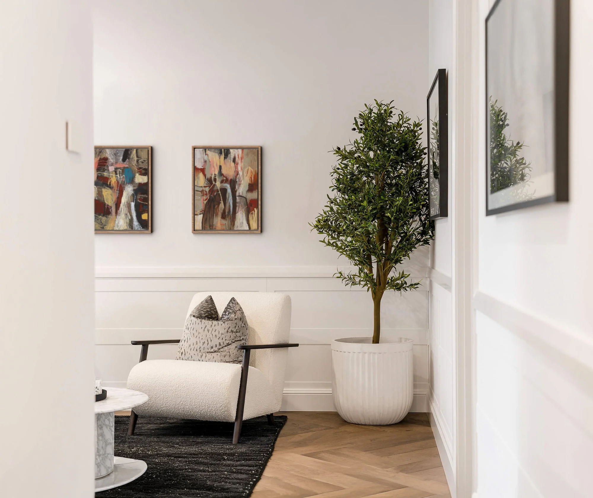 Interior space with two framed abstract paintings on a light-colored wall, a tall potted plant in a white planter, a white upholstered chair with a throw pillow, a white marble-top table, and a dark rug on wooden flooring.