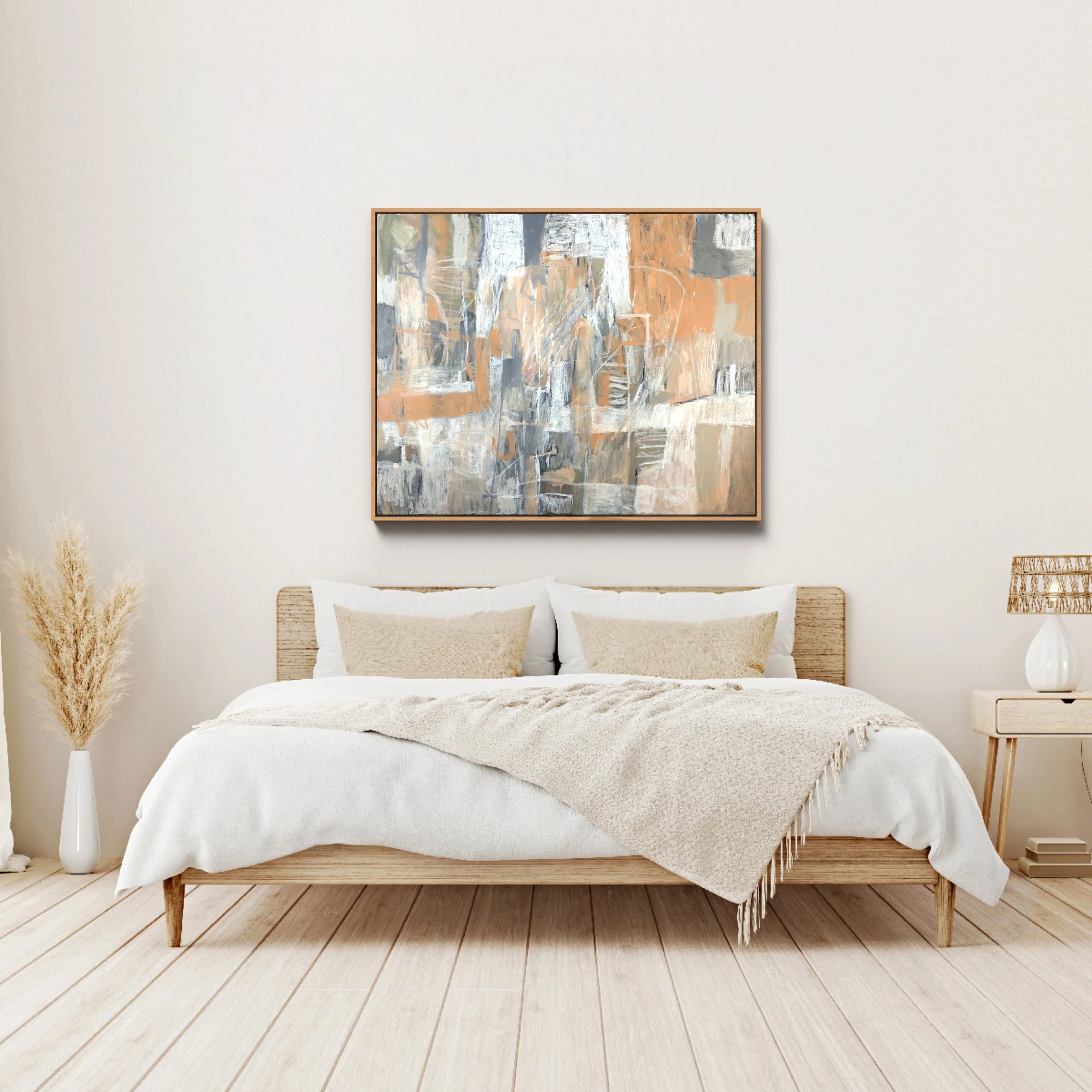 A minimalist bedroom with a wooden bed frame, white bedding, beige pillows, a framed abstract painting hanging above the bed, a woven side table, a tall white vase with dried pampas grass, and a wooden floor.