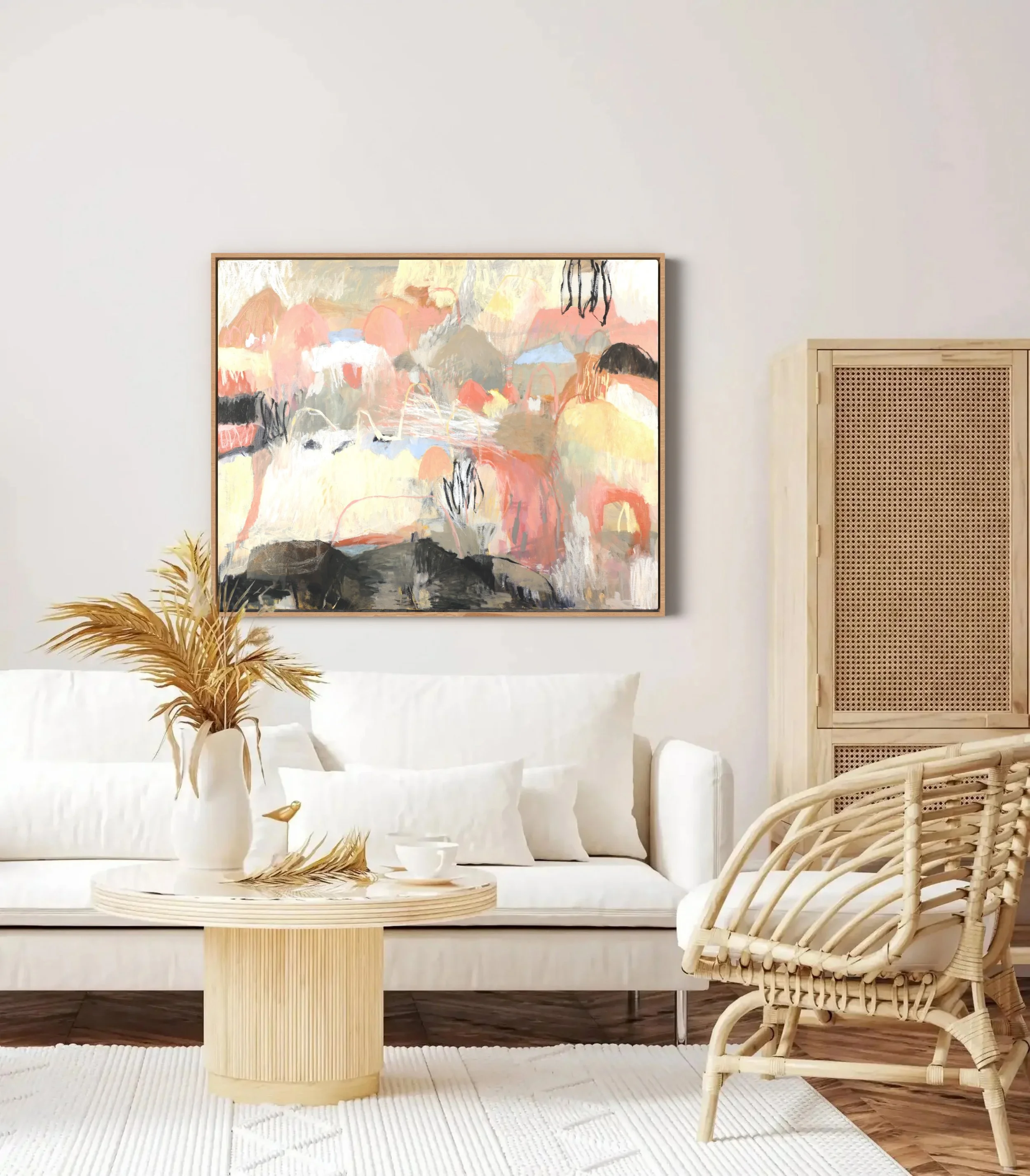 A living room with a white sofa, a round wooden coffee table with a vase of dried plants, a framed abstract painting on the wall, a cream-colored woven rattan chair, a white textured rug, and a beige textured wall panel.