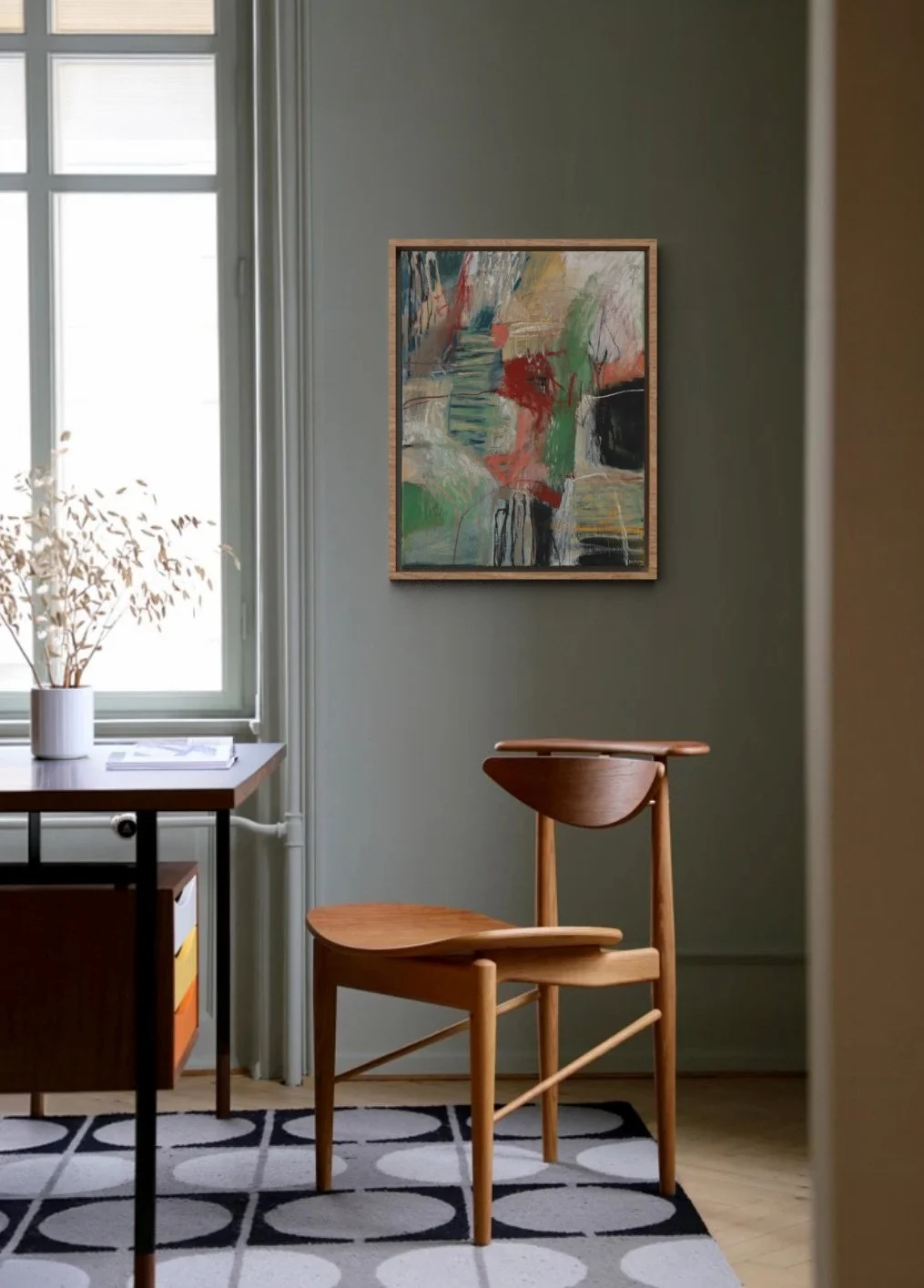 A corner of a room featuring a wooden chair with a curved backrest, a table with a white vase holding dried branches near a window, an abstract painting on the wall, and a patterned rug on the floor.