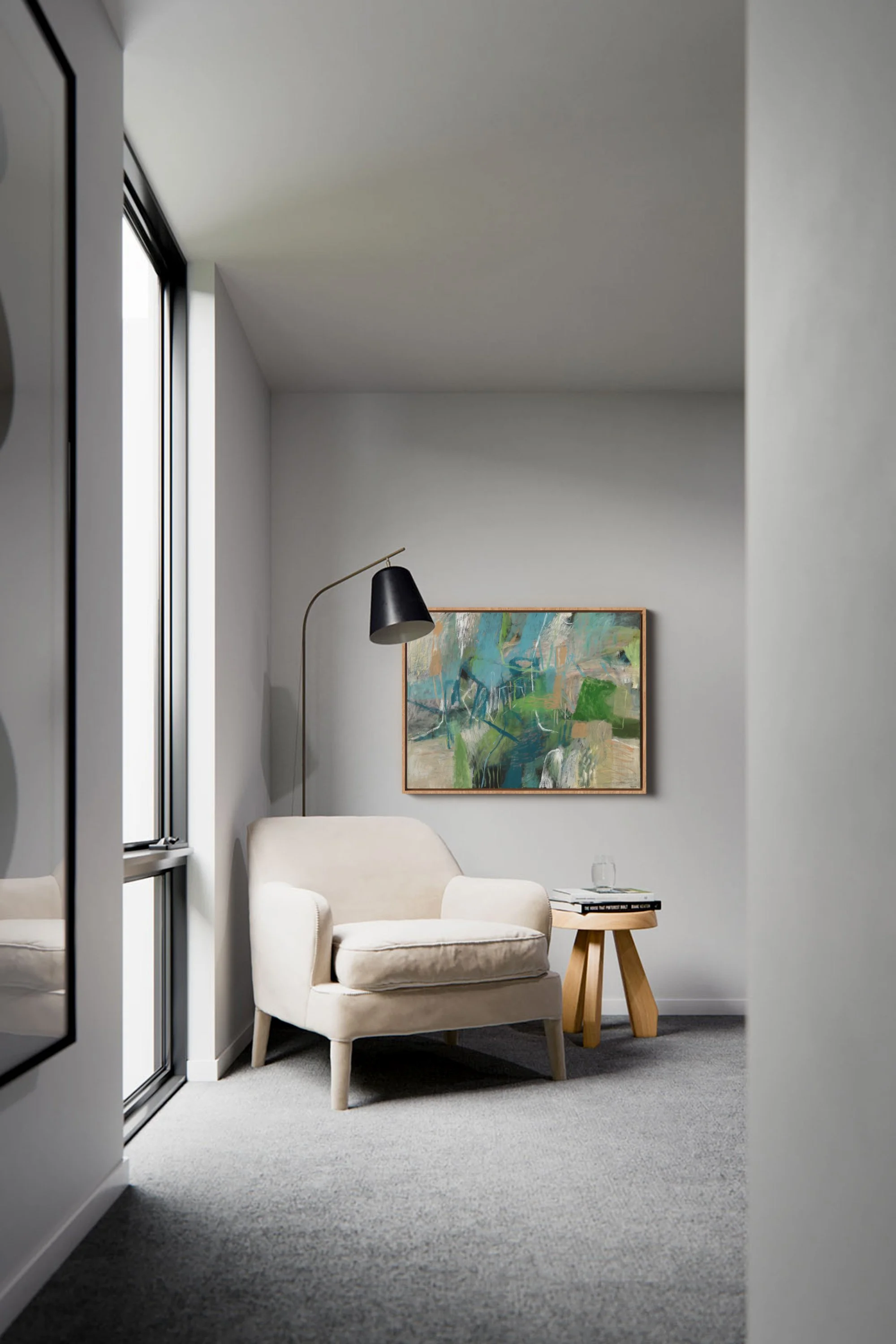 Cozy living room with beige armchair, small wooden table holding a glass of water and a book, colorful abstract painting on the wall, black floor lamp, gray walls, large window letting in natural light.