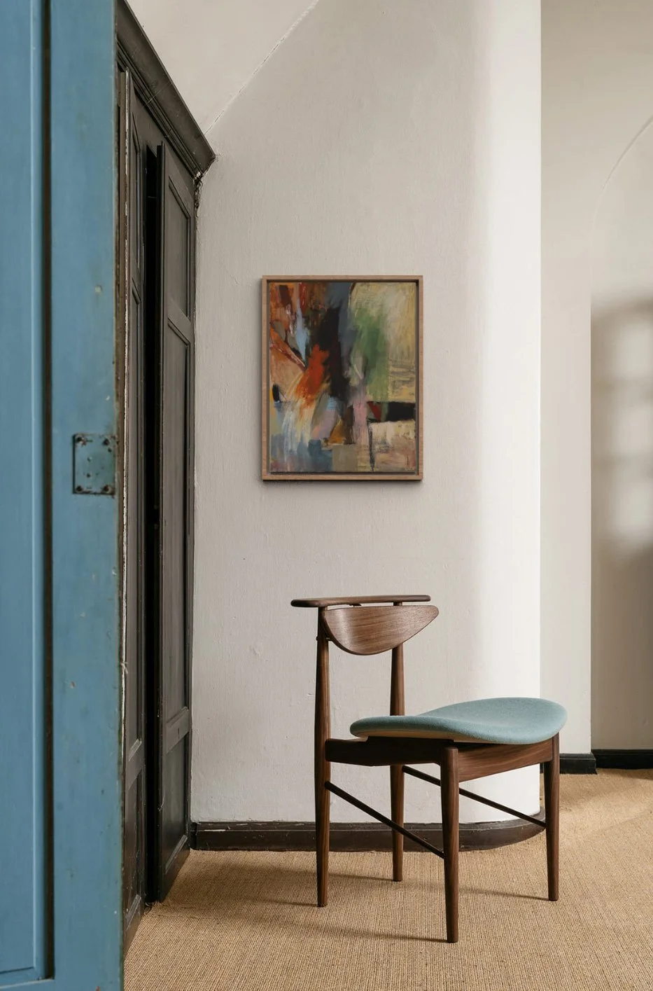 A wooden chair with light blue upholstery, positioned against a white textured wall in a room with beige carpet. A colorful abstract painting hangs on the wall above the chair, and a black door frame is partially visible on the left.