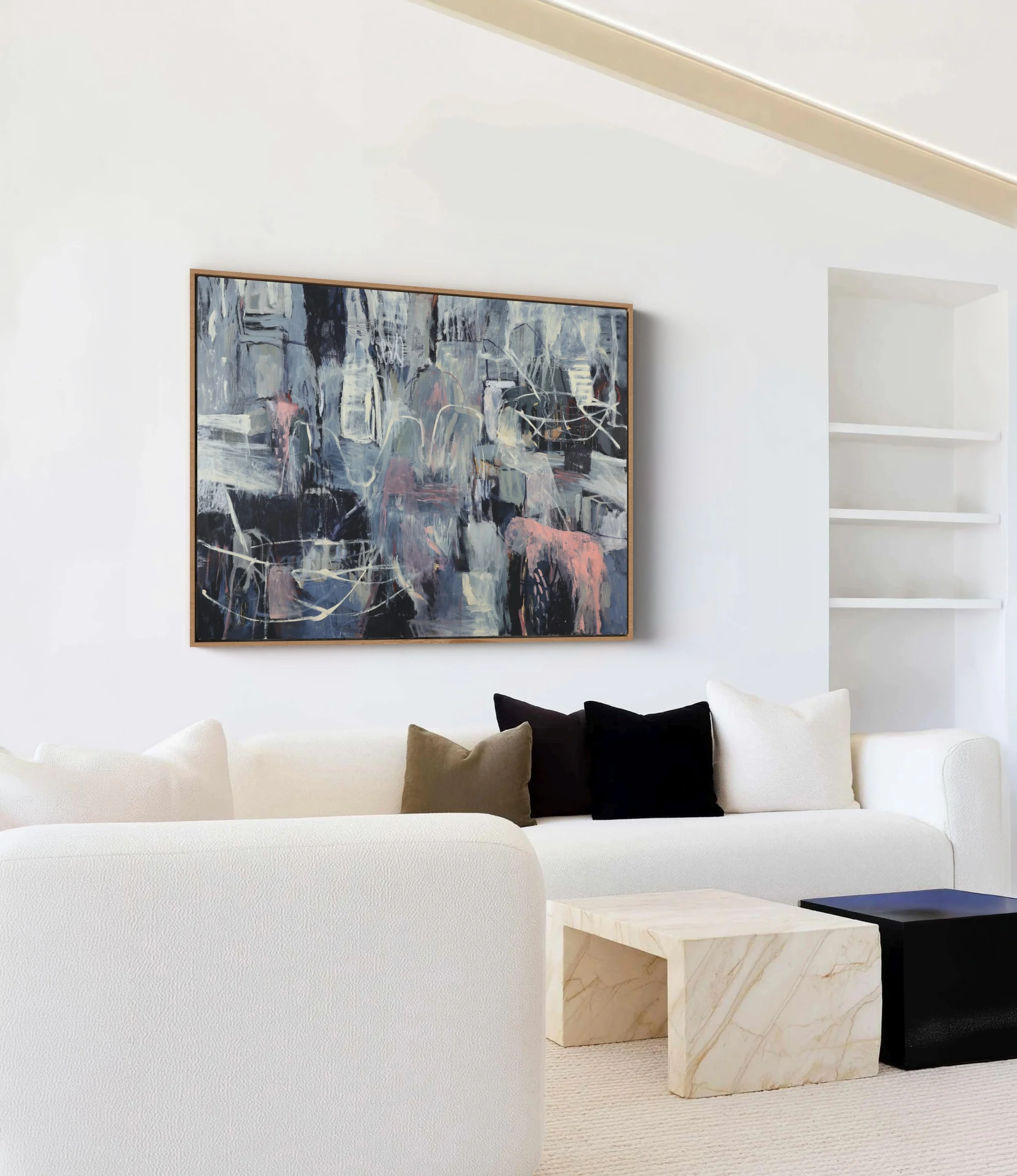 A minimalist living room with a white curved sofa, black and beige pillows, a marble side table, an abstract painting on the wall, and built-in white shelves on the wall.