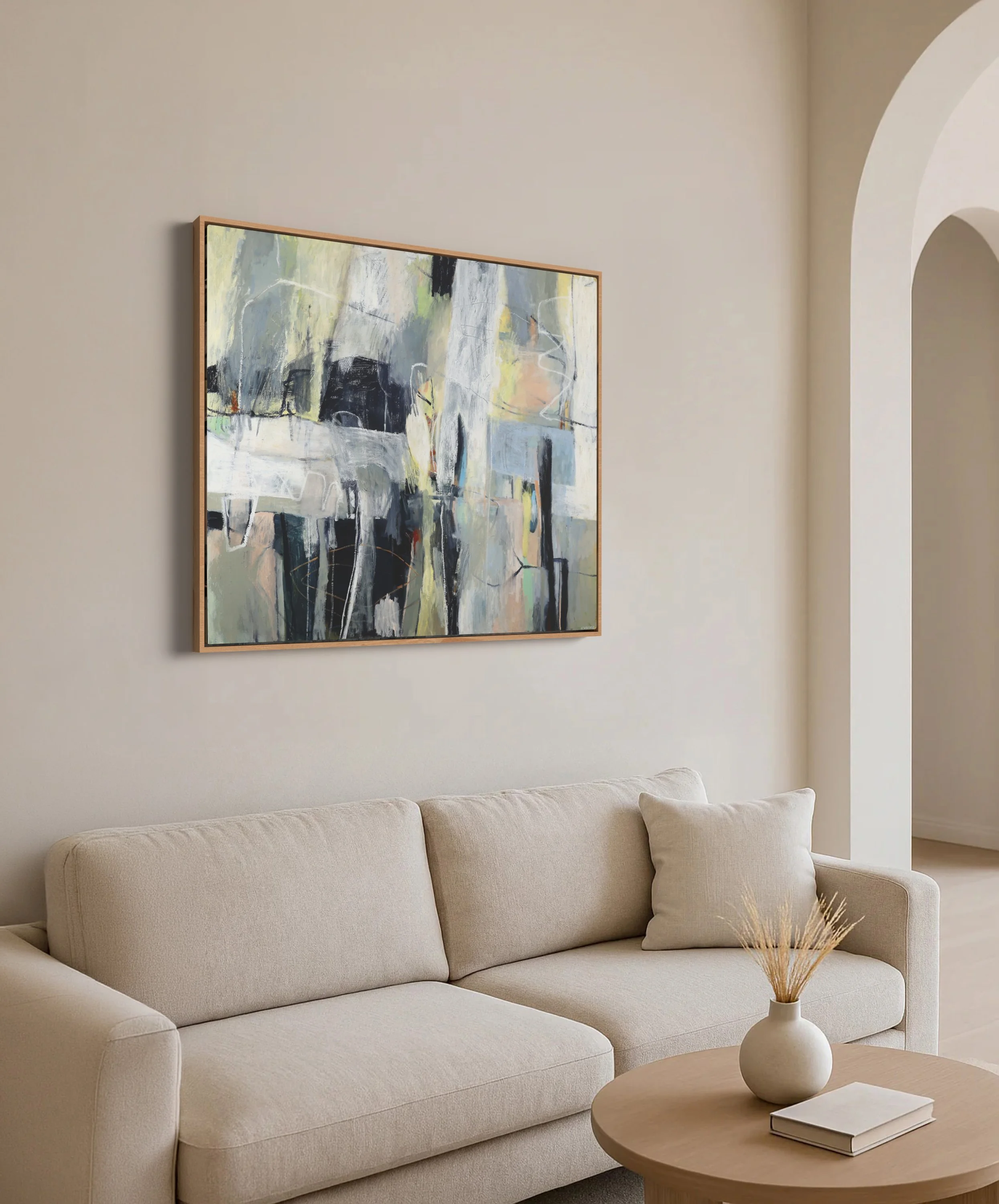 A beige sofa with plush cushions in a modern living room. A round wooden coffee table with a white vase holding dried grass and a closed book is in front. An abstract painting with black, white, and pastel colors hangs on the wall. The room has neutr