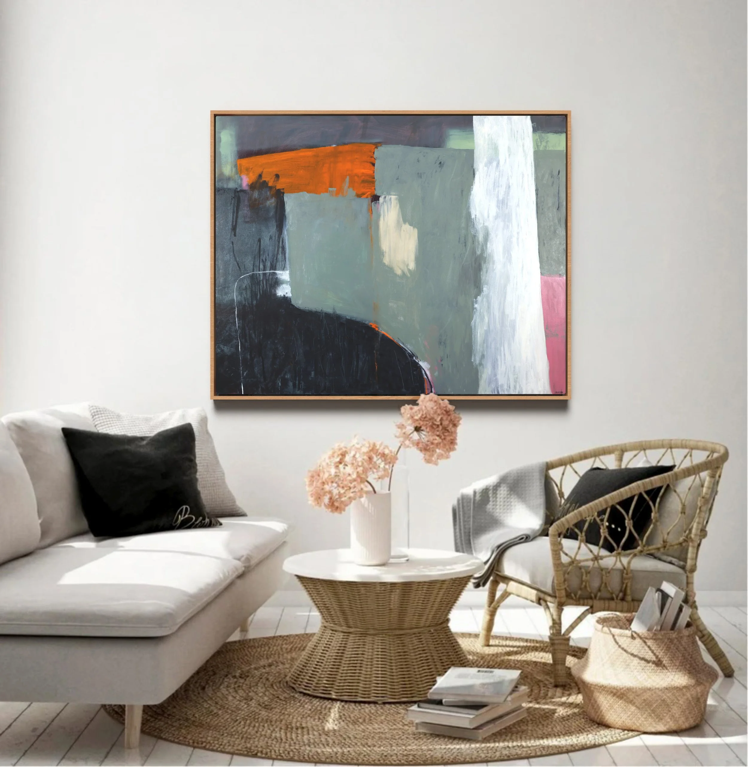 A cozy living room with white walls and a large abstract painting in shades of black, gray, orange, white, and pink hanging above a white sofa. The sofa has a black pillow and a blanket. In front of the sofa is a round wicker coffee table with a bouq