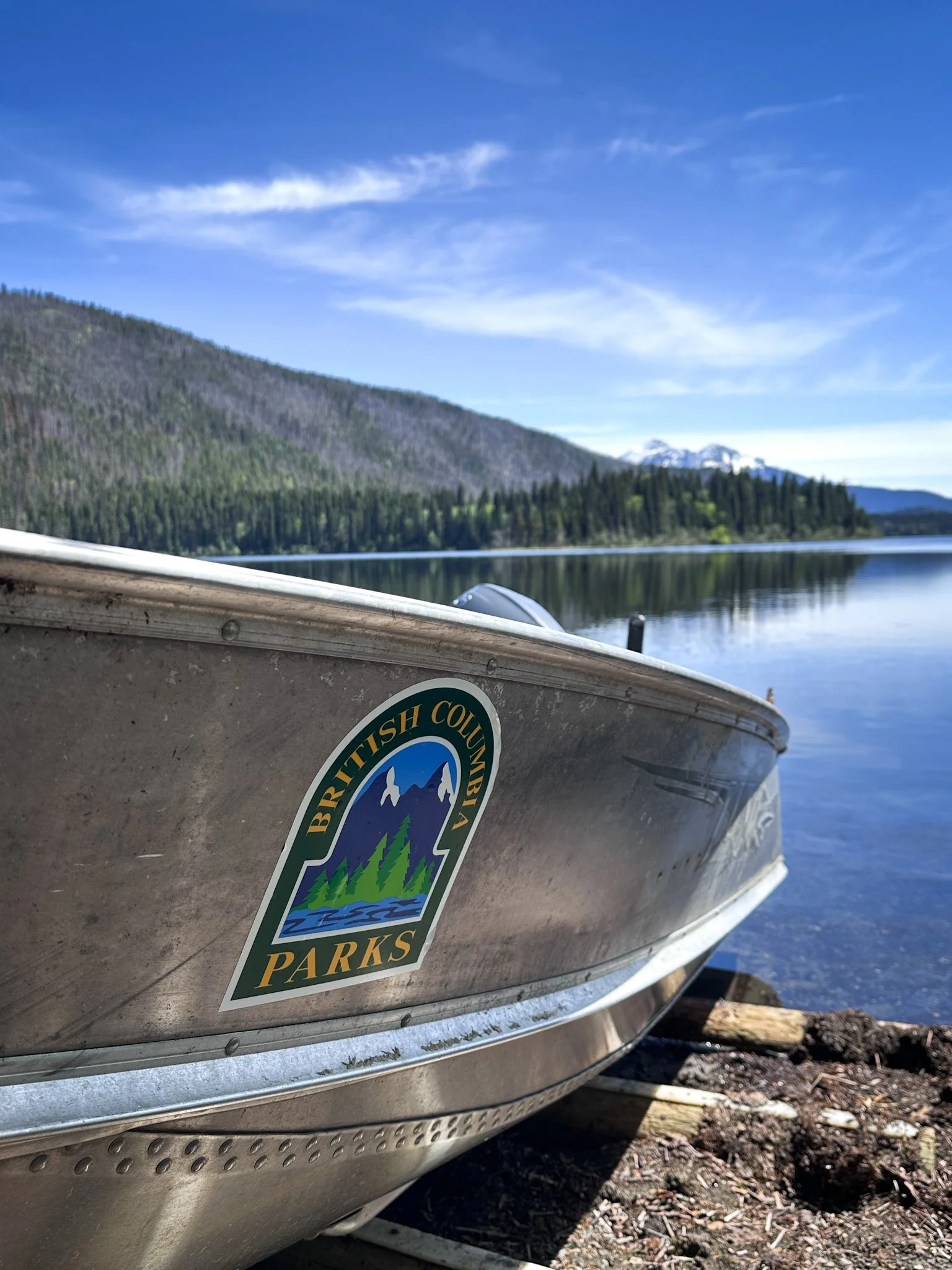 Close-up of a silver aluminum boat with a British Columbia Parks sticker, beside a calm lake with forested mountains and a clear blue sky in the background on the Bowron Lake Circuit