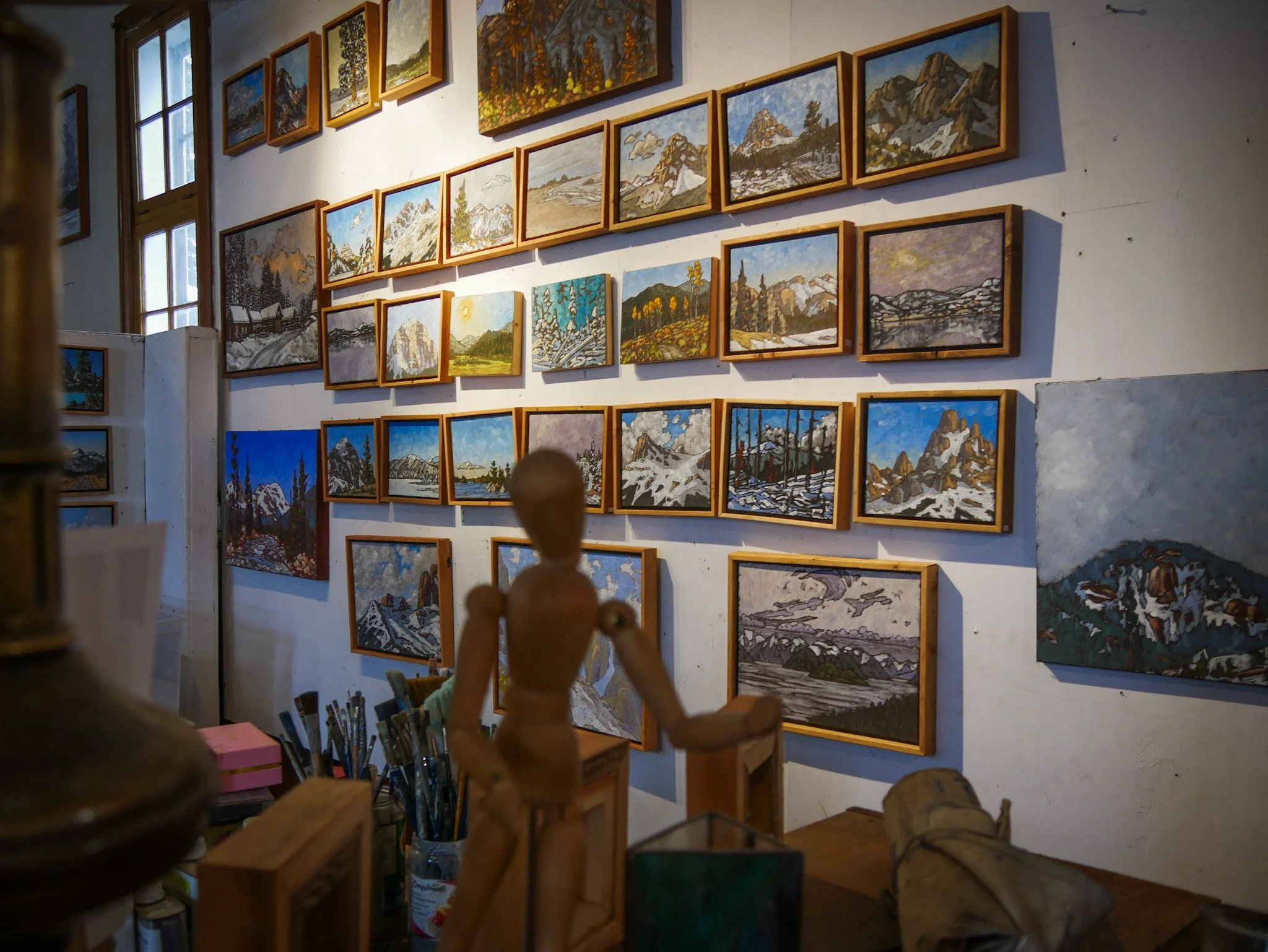 Art gallery wall at St George Studio displaying multiple landscape paintings of Peter Corbett in wooden frames, with various mountain, forest, and natural scenery, art supplies including brushes and paints on a table in the foreground.