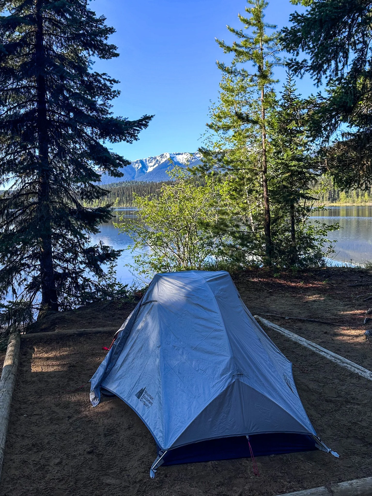 A blue camping tent set up near a calm lake surrounded by trees, with snow-capped mountains of the Cariboo Mt Range in the background under a clear blue sky on the Bowron Lake Circuit