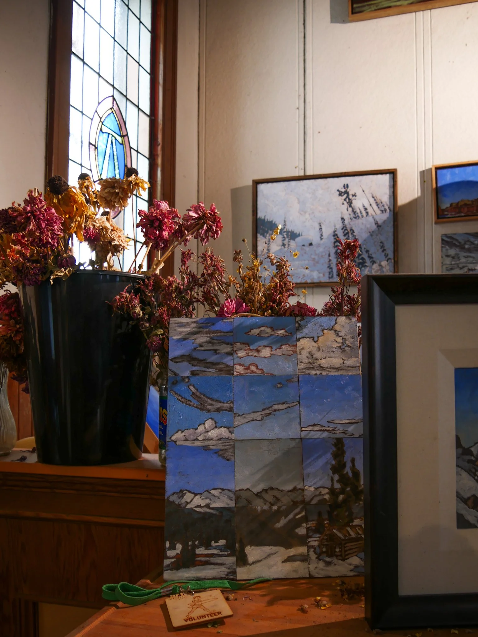 St George Studio with a stained glass window, framed landscape paintings on the wall, a tall black vase with dried pink and yellow flowers, a wooden table with a wooden volunteer badge, and a tiled wall behind the artworks of Peter Corbett