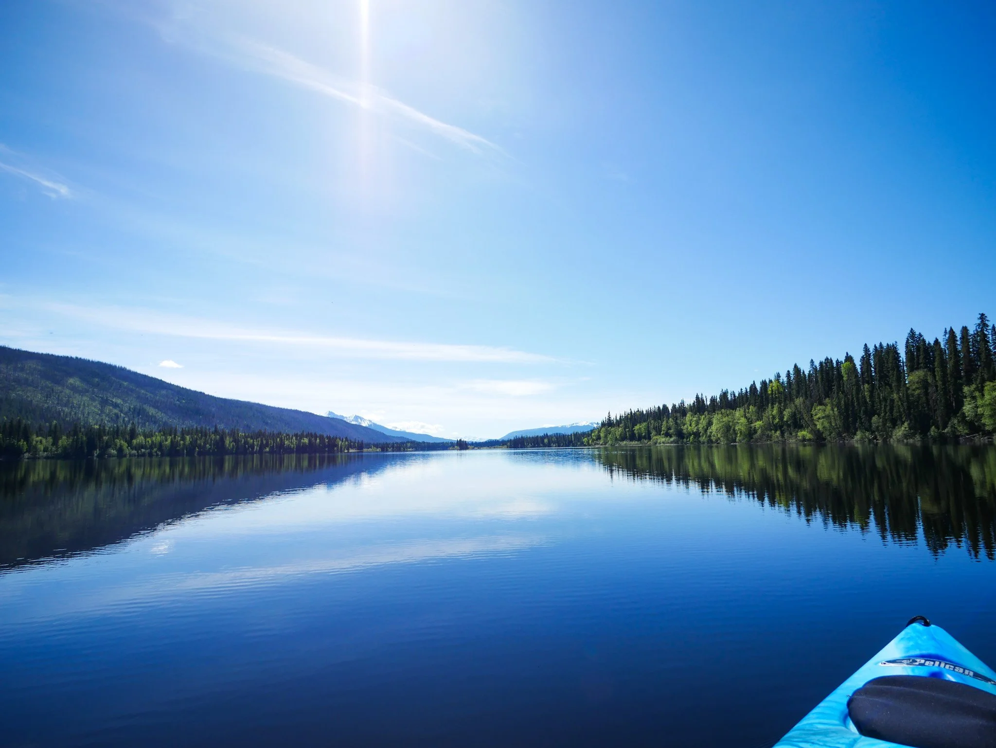 A peaceful lake surrounded by green forested hills with snow-capped mountains in the distance under a clear blue sky, as seen from a kayak  on the Bowron Lake Circuit