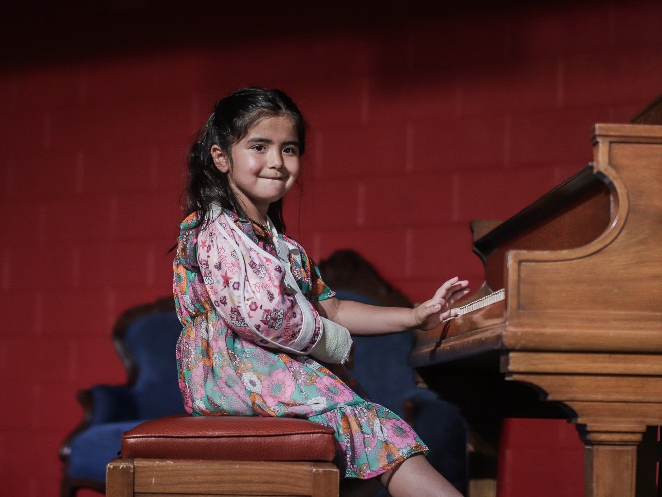 Young girl with long dark hair playing the piano, sitting on a red cushioned stool, wearing a colorful patterned dress, in front of a red brick wall.