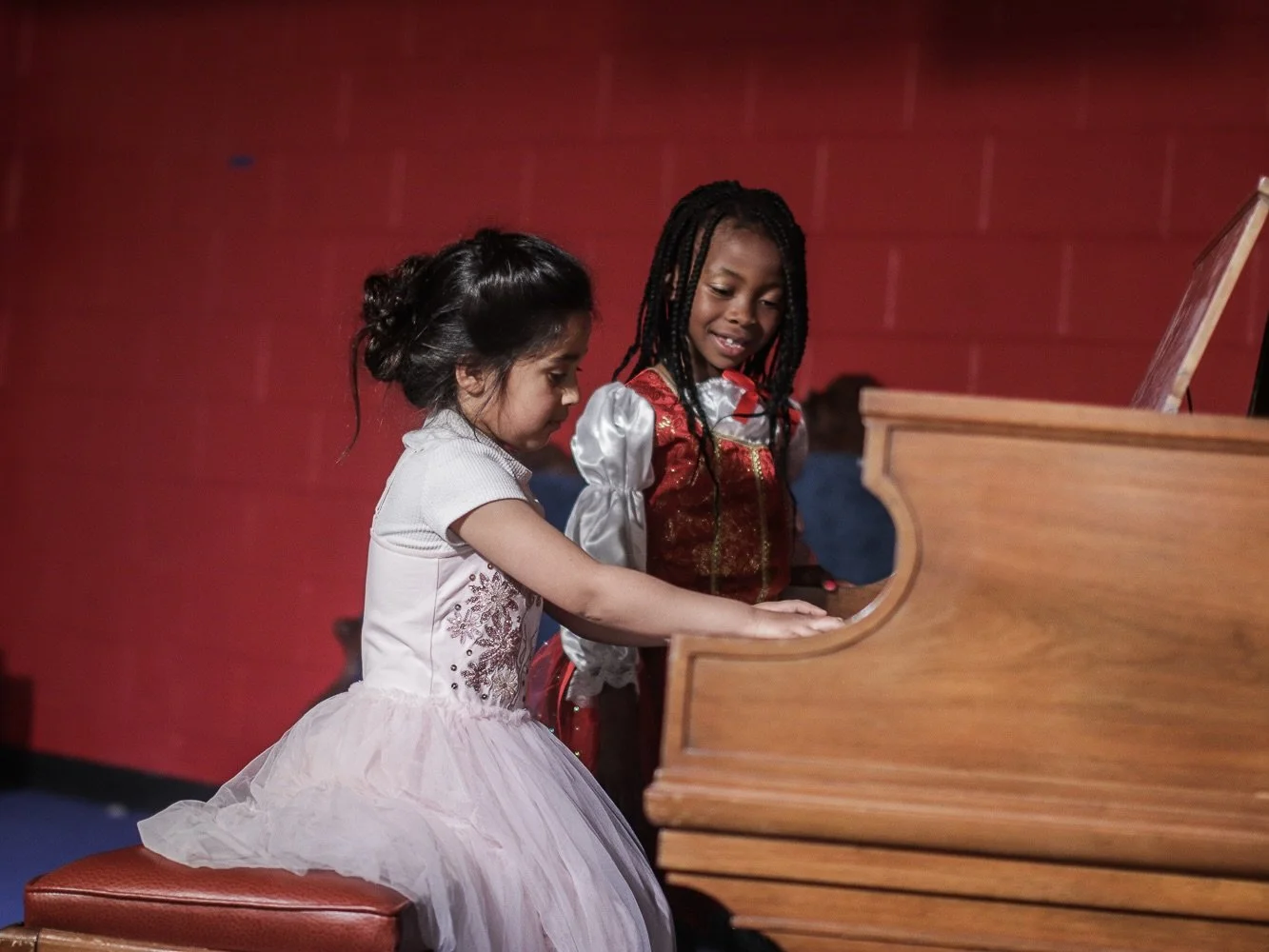 Two young girls playing the piano together, one sitting on a stool and the other standing, in a room with a red wall.