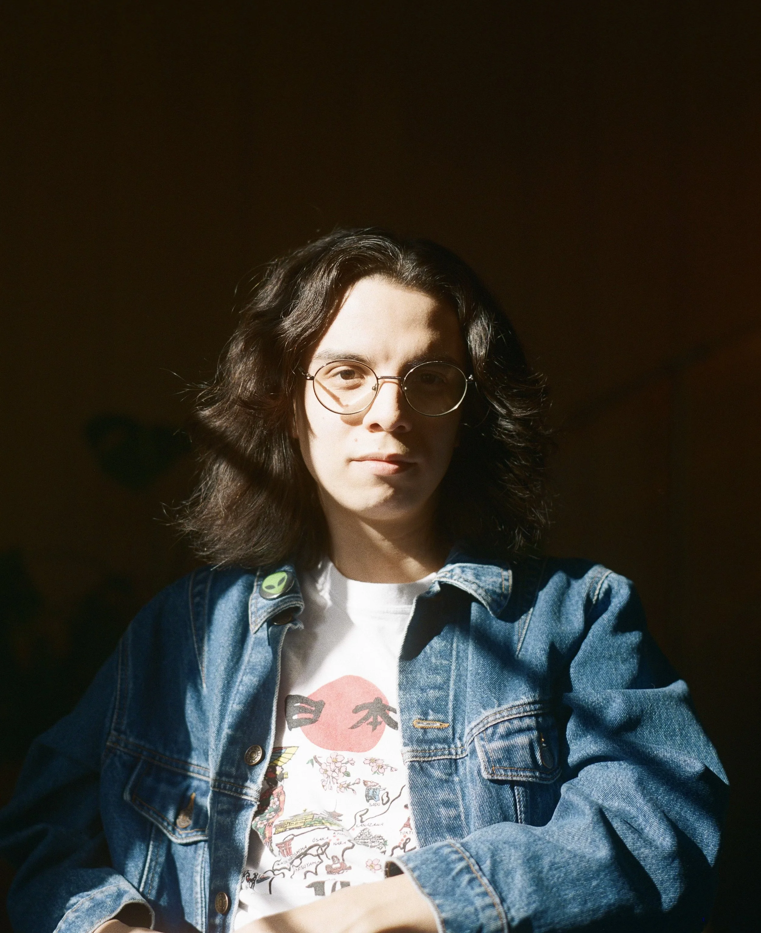 A young man with shoulder length dark brown hair wearing glasses and a denim jacket with a pleasant soft smile.
