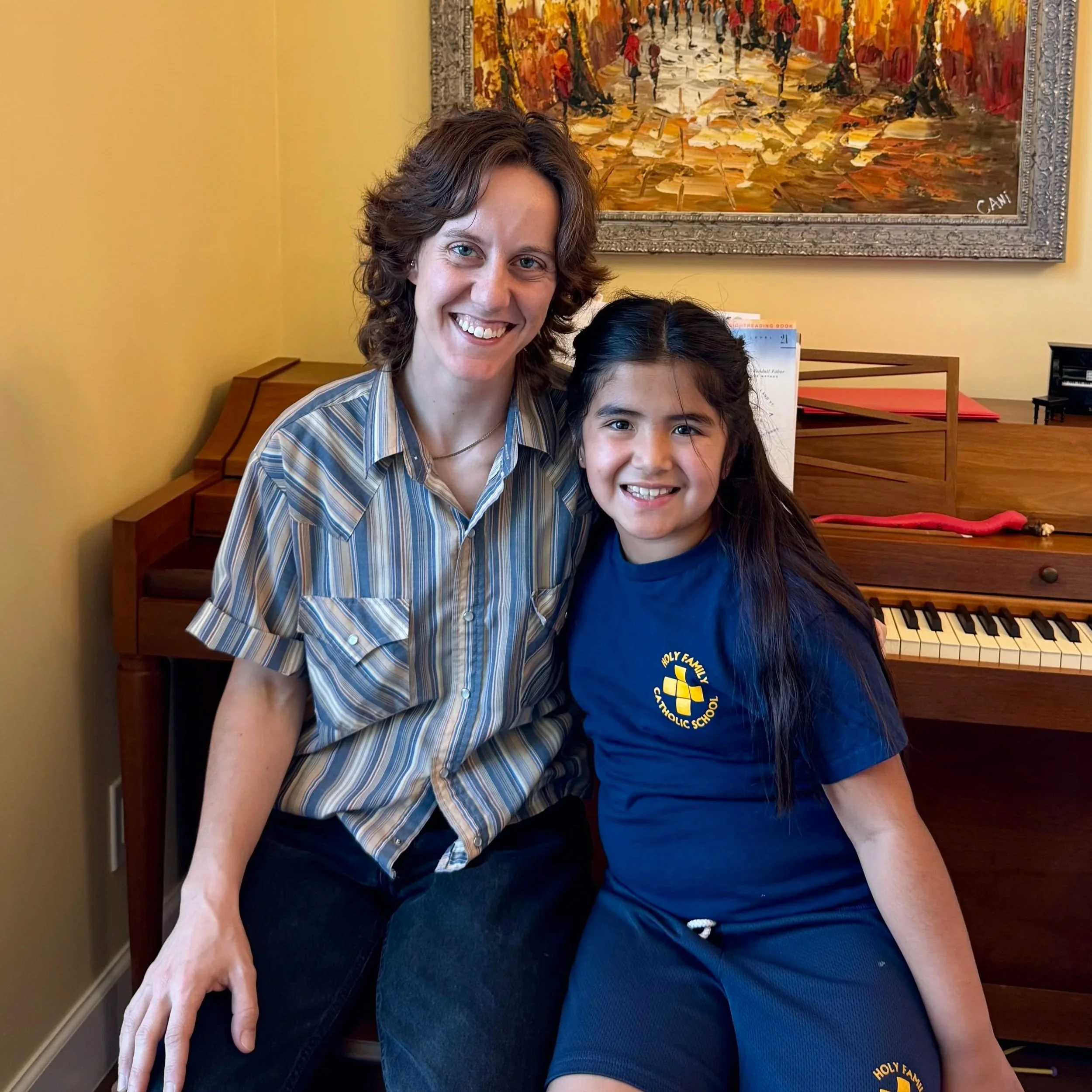 A woman and a young girl sitting together in front of a piano, both smiling at the camera. The woman is wearing a striped shirt, and the girl is wearing a blue shirt with a school logo, in a room with a yellow wall and a framed painting background.