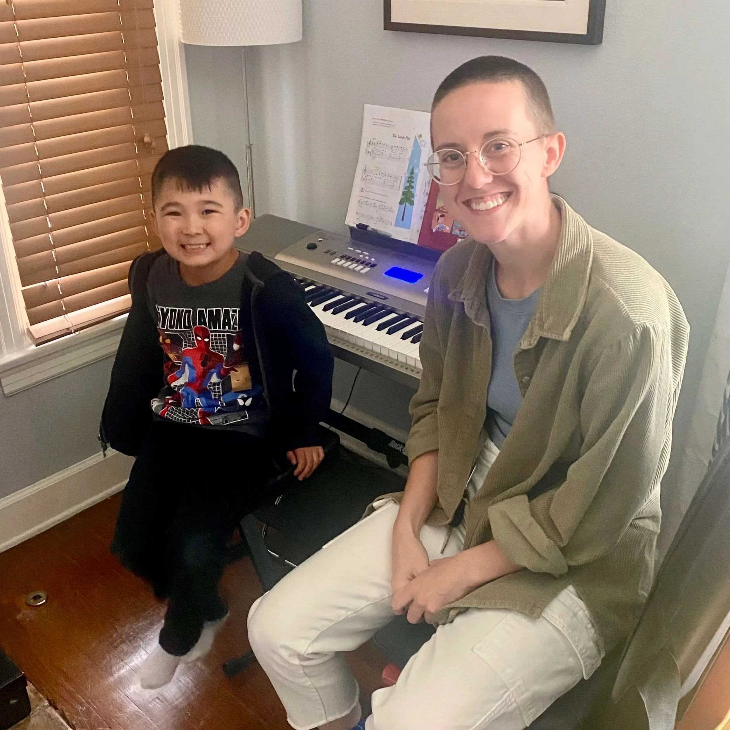 A young boy and an adult woman sitting indoors near a digital keyboard. The boy is smiling and wearing a Spider-Man T-shirt with a black hoodie. The woman is smiling, wearing glasses, a beige jacket, and white pants. The room has wooden floors, a window with brown blinds, and some music sheets on the keyboard.