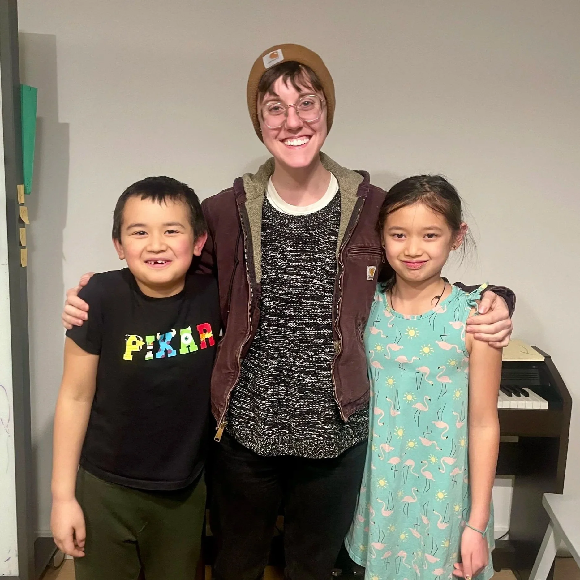 Smiling woman with glasses and short brown hair, wearing a beanie, standing with her arms around a young boy and girl, in front of a plain wall and a small keyboard.