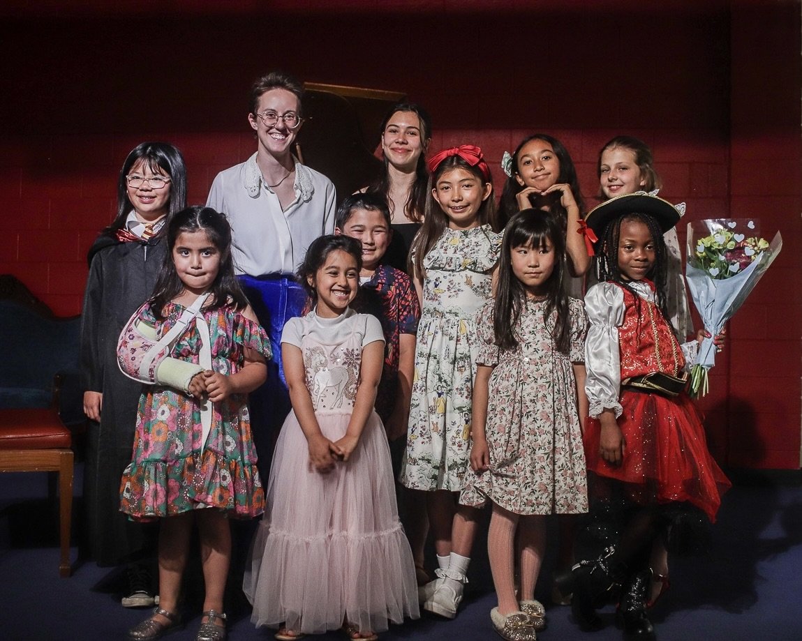 A group of young girls and two adult women posing together on a stage, some girls wearing costumes and holding flowers, with a red wall in the background.