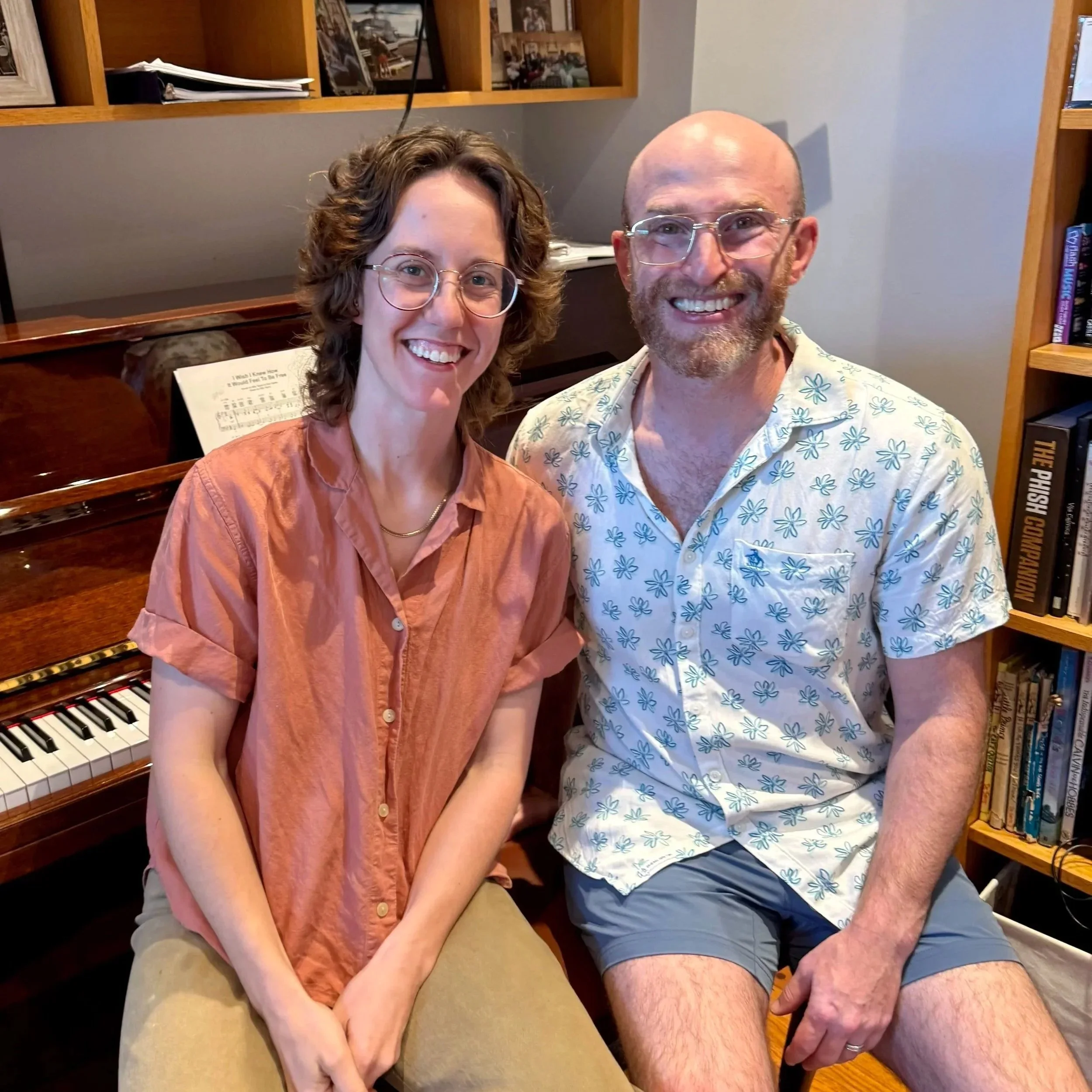 A woman and a man sitting indoors, smiling at the camera. The woman is wearing glasses and an orange button-up shirt, and has curly brown hair. The man is bald, also wearing glasses, with a beard, and is dressed in a light-colored Hawaiian shirt with blue floral patterns and shorts. Behind them is a piano with some sheet music and a bookshelf.