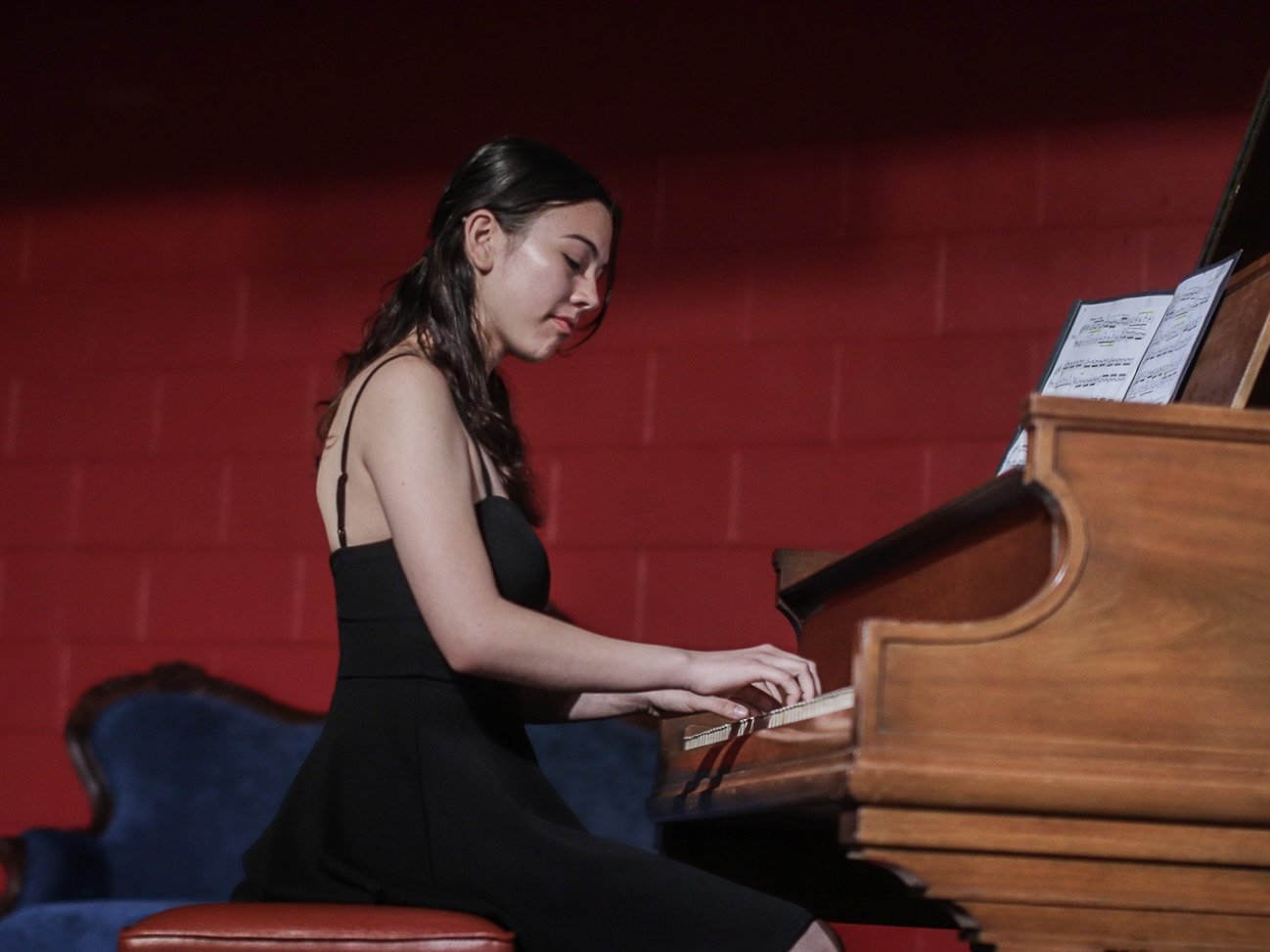 Young woman playing piano on stage with sheet music, red wall background.
