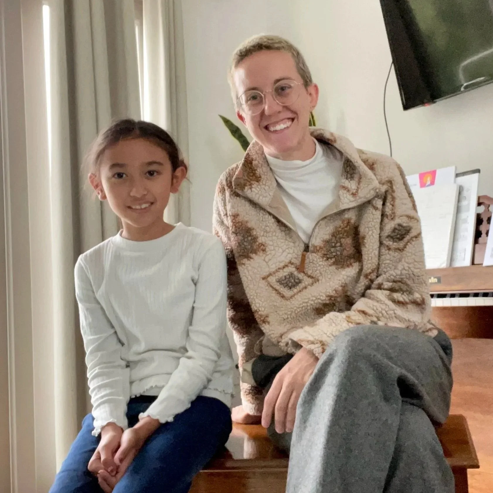 A young girl and an adult woman sitting together on a wooden bench in a room with cream curtains, a TV, and a piano in the background.