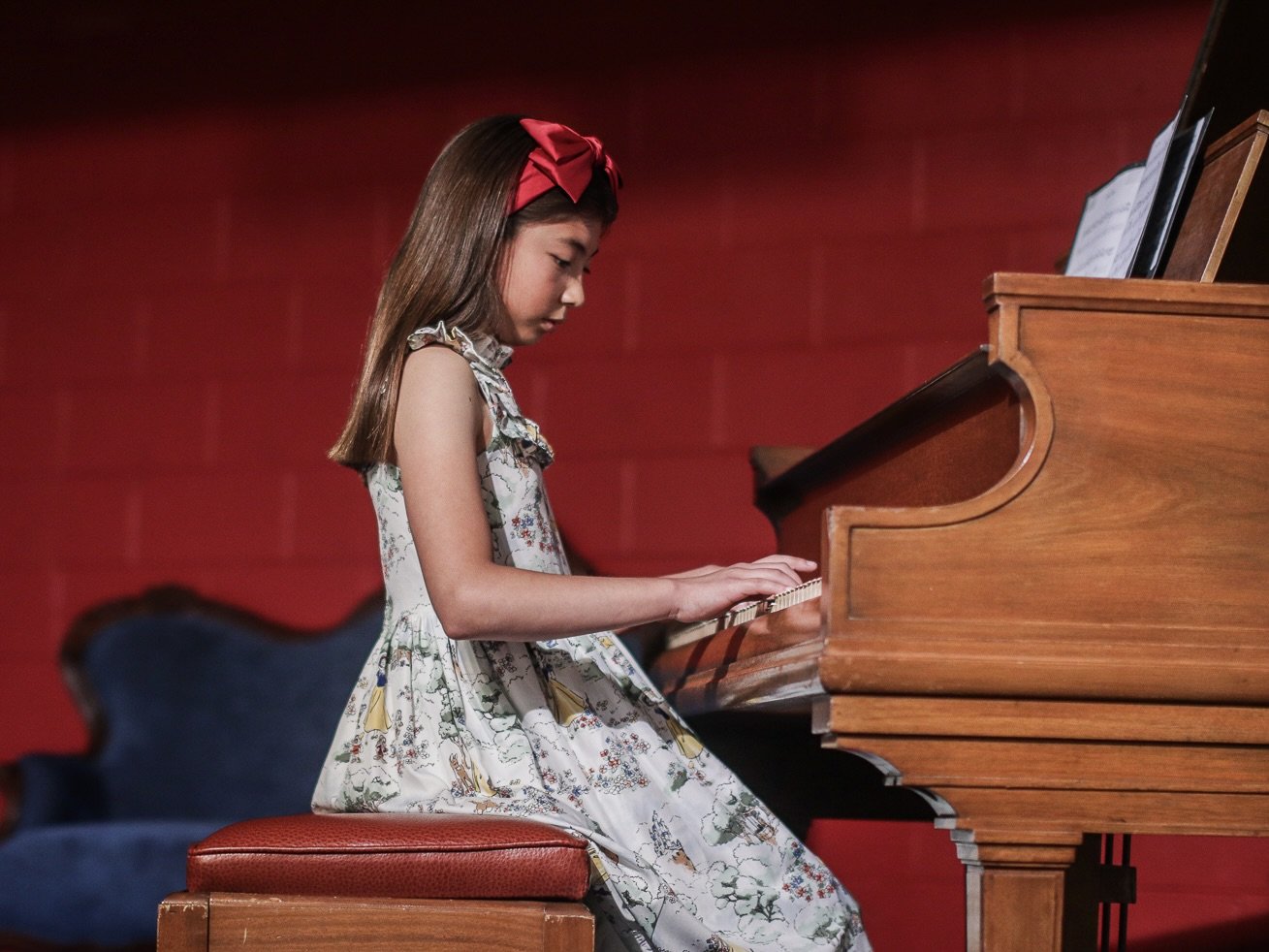 A young girl with long hair wearing a floral dress and a red headband playing a wooden piano.
