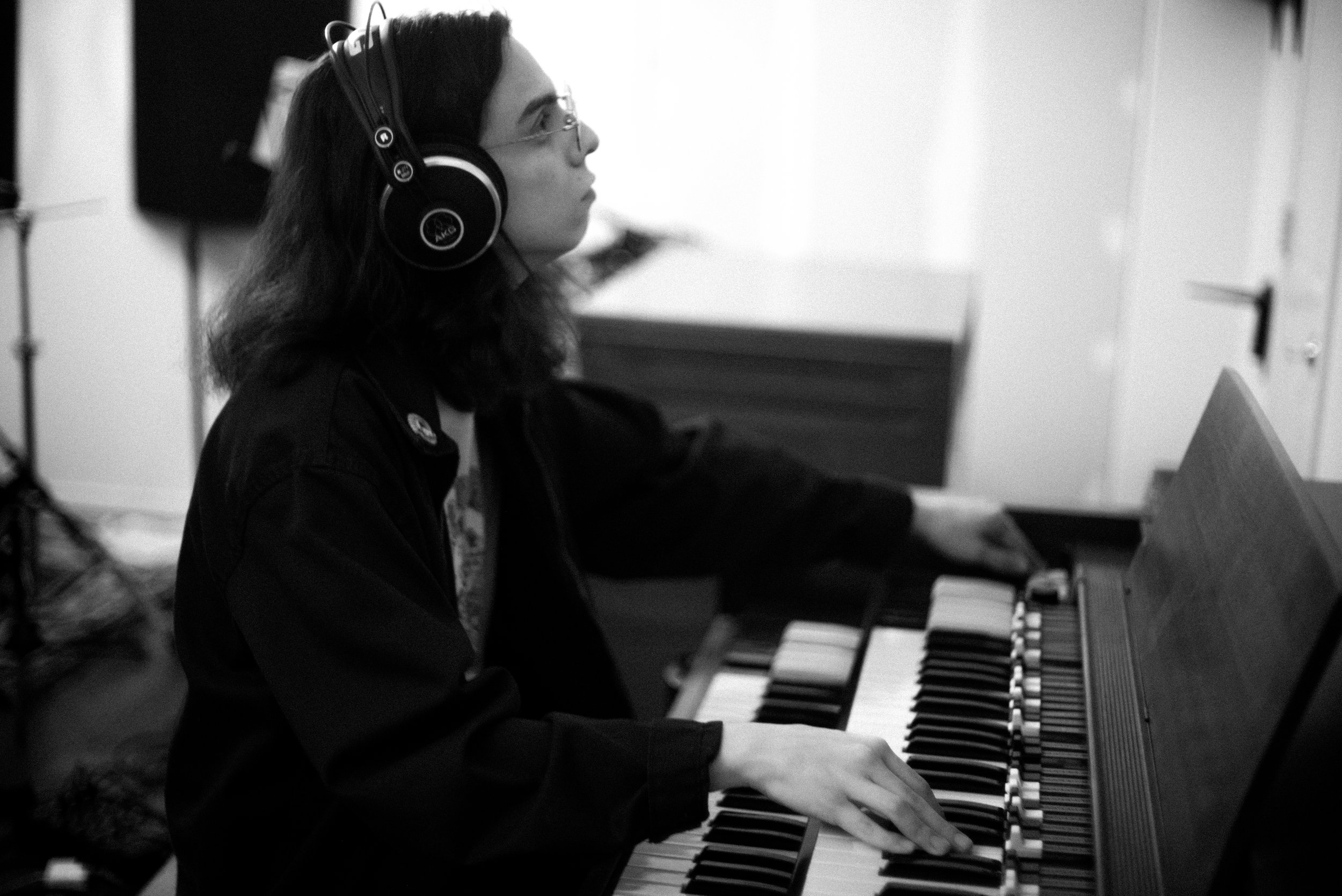 A young man with shoulder length dark brown hair wearing a glasses and black jacket and studio headphones playing a keyboard organ in a music studio.