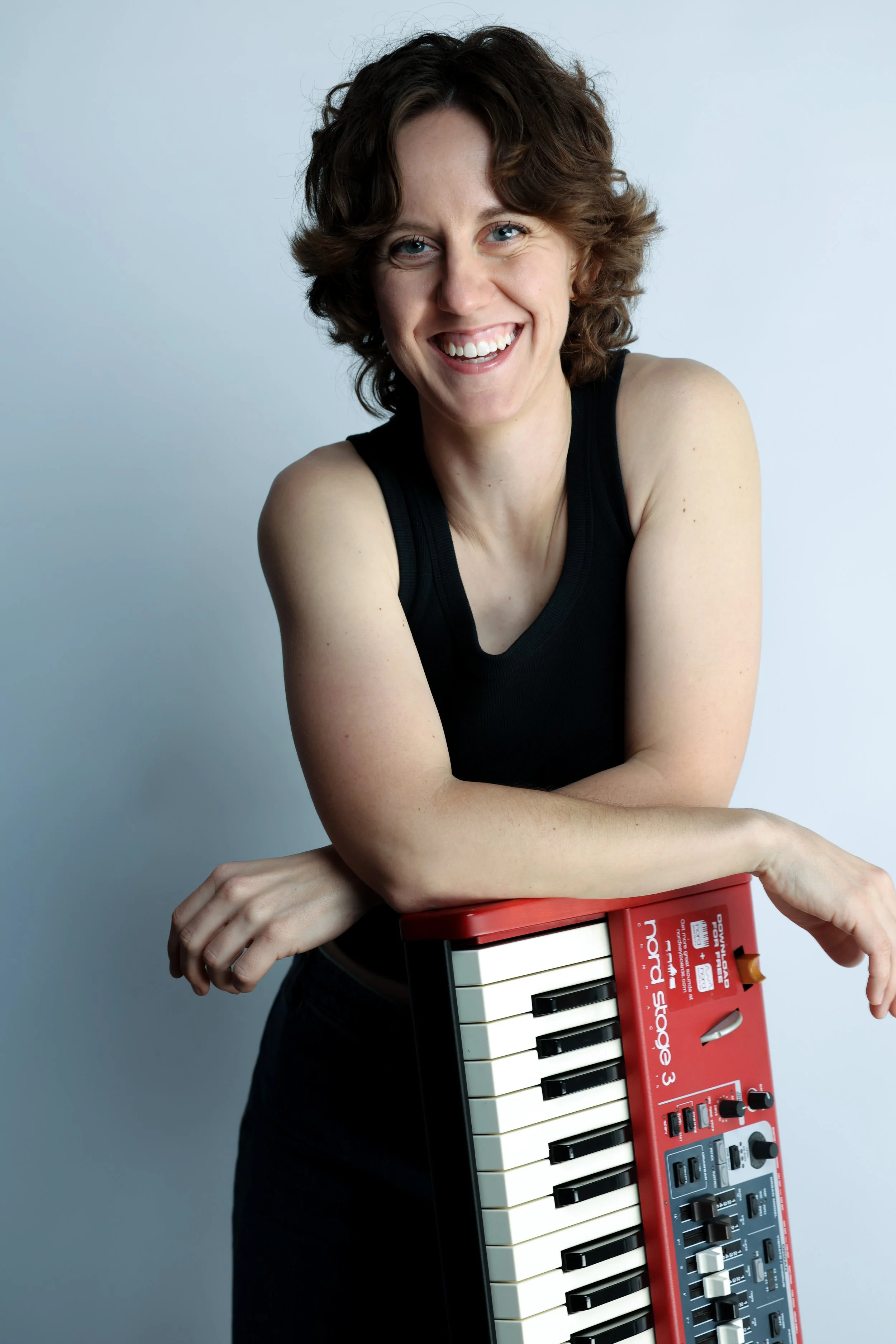A woman with curly brown hair wearing a black sleeveless top holding a red musical keyboard and smiling.