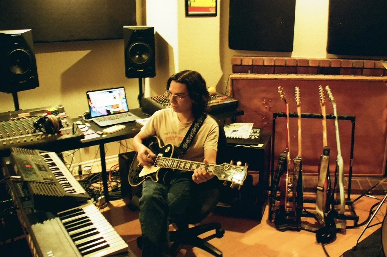A young man with shoulder length dark brown hair wearing a glasses and a t-shirt playing guitar in a music studio.