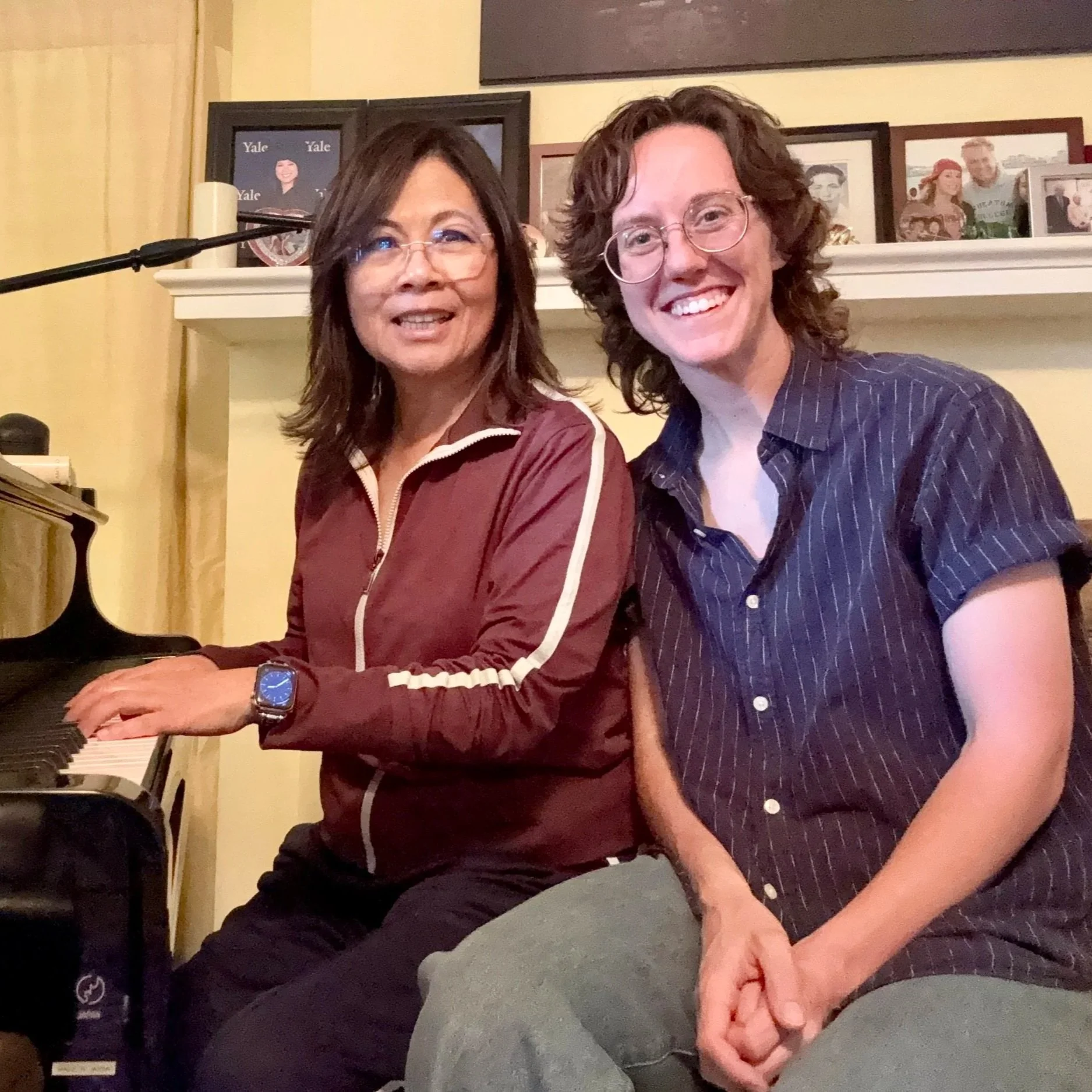 A woman playing the piano next to a person sitting on a bench, in a room decorated with framed photos on a white shelf.