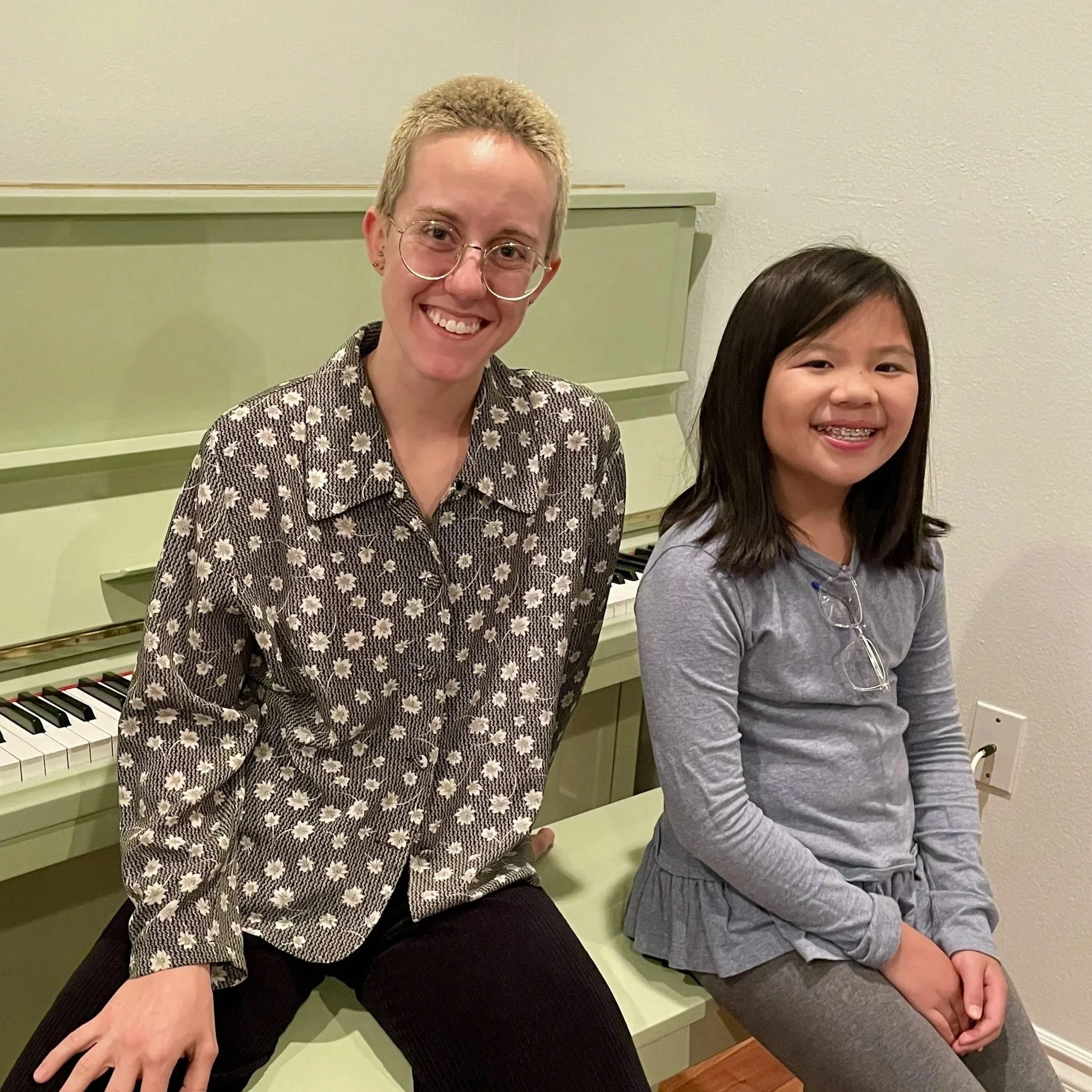 Two smiling females, one adult and one child, sitting near a green piano in a room with beige walls.