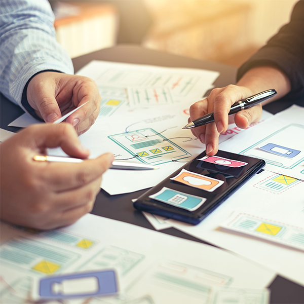 Two people working on mobile app design sketches and wireframes at a table, with sketches, papers, and a smartphone displaying app interface elements.