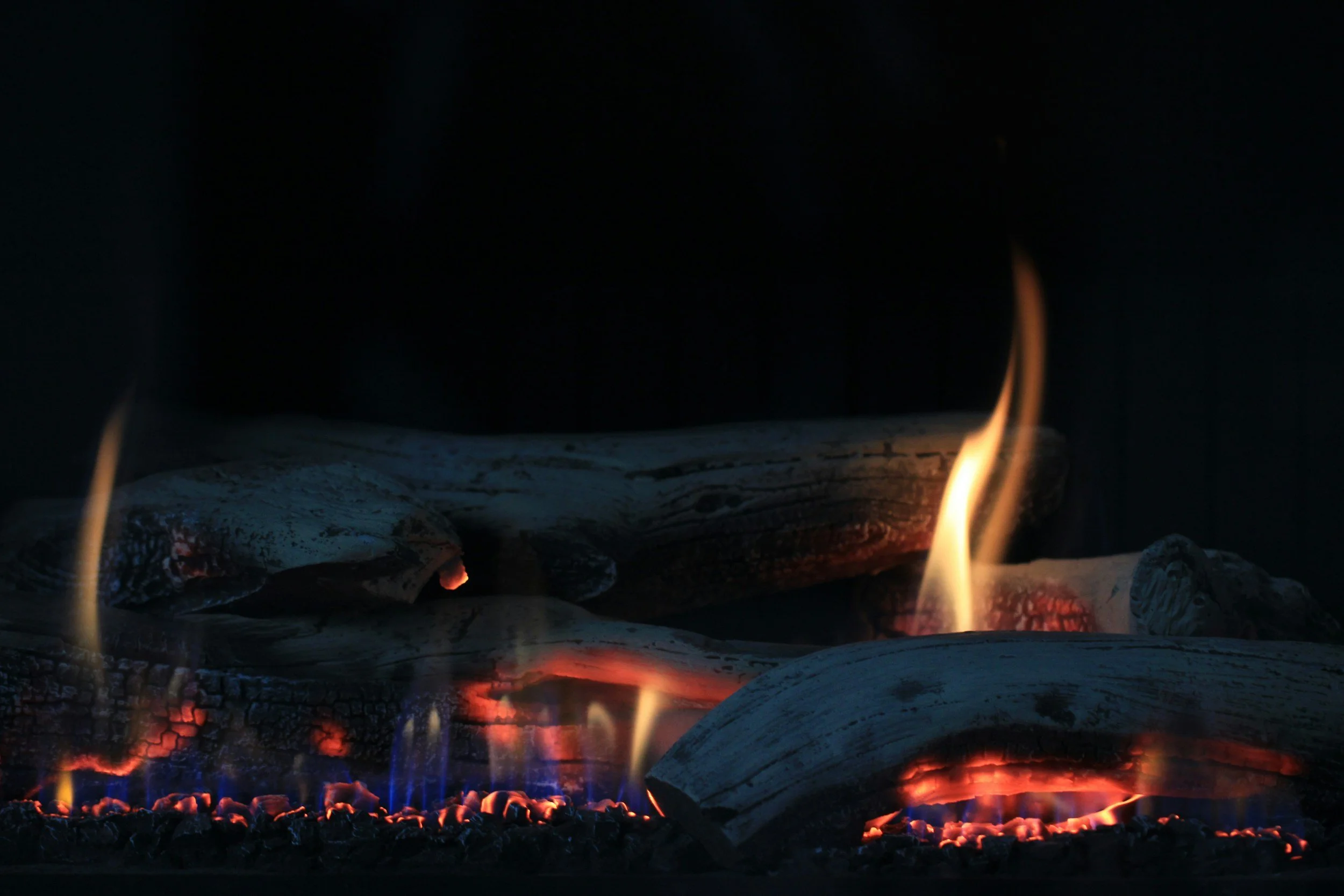 A close-up of a gas fireplace with burning logs, orange flames, and glowing embers in a dark background.