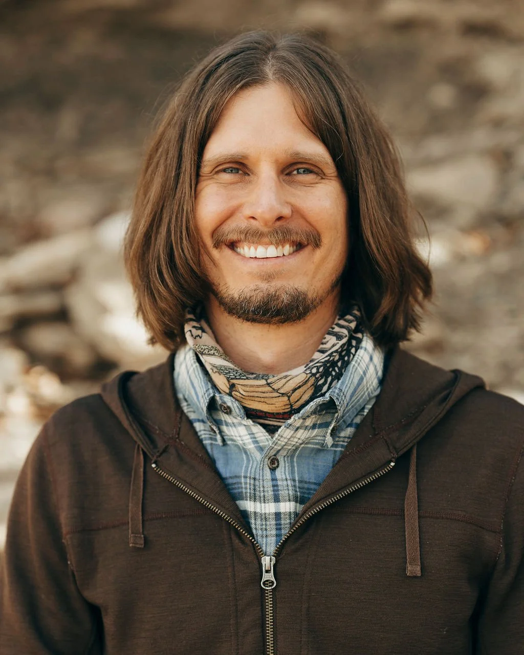 A smiling man with shoulder-length brown hair, a mustache, and a beard. He's wearing a dark hoodie, a plaid shirt, and a patterned bandana around his neck, outdoors with a blurred natural background.